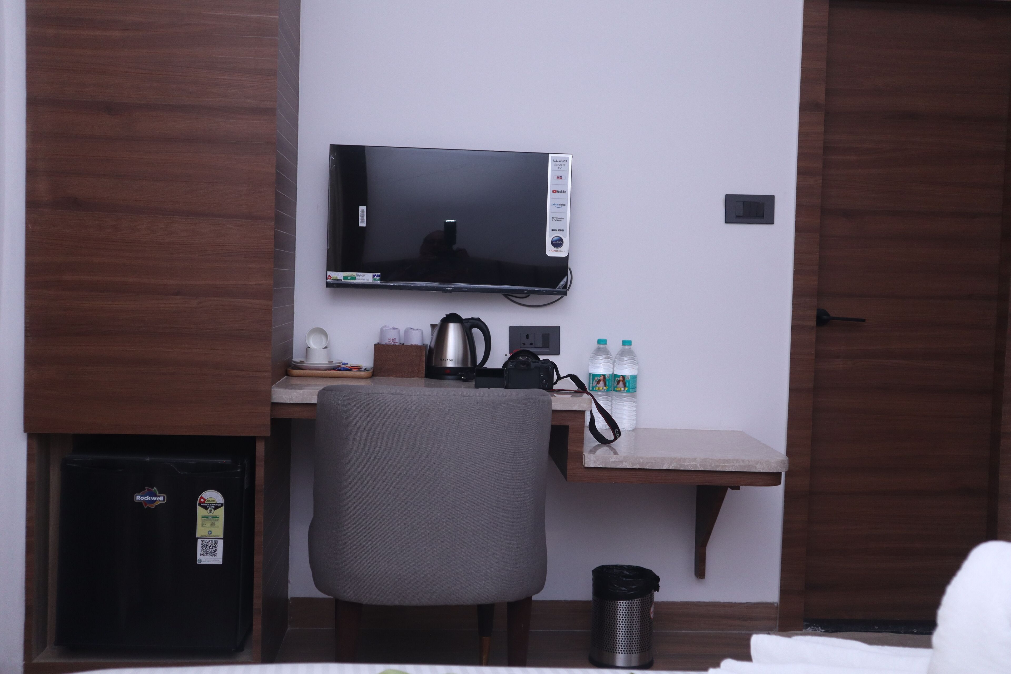 Habitación de hotel con cama, escritorio, monitor de computadora, silla, cafetera y refrigerador.