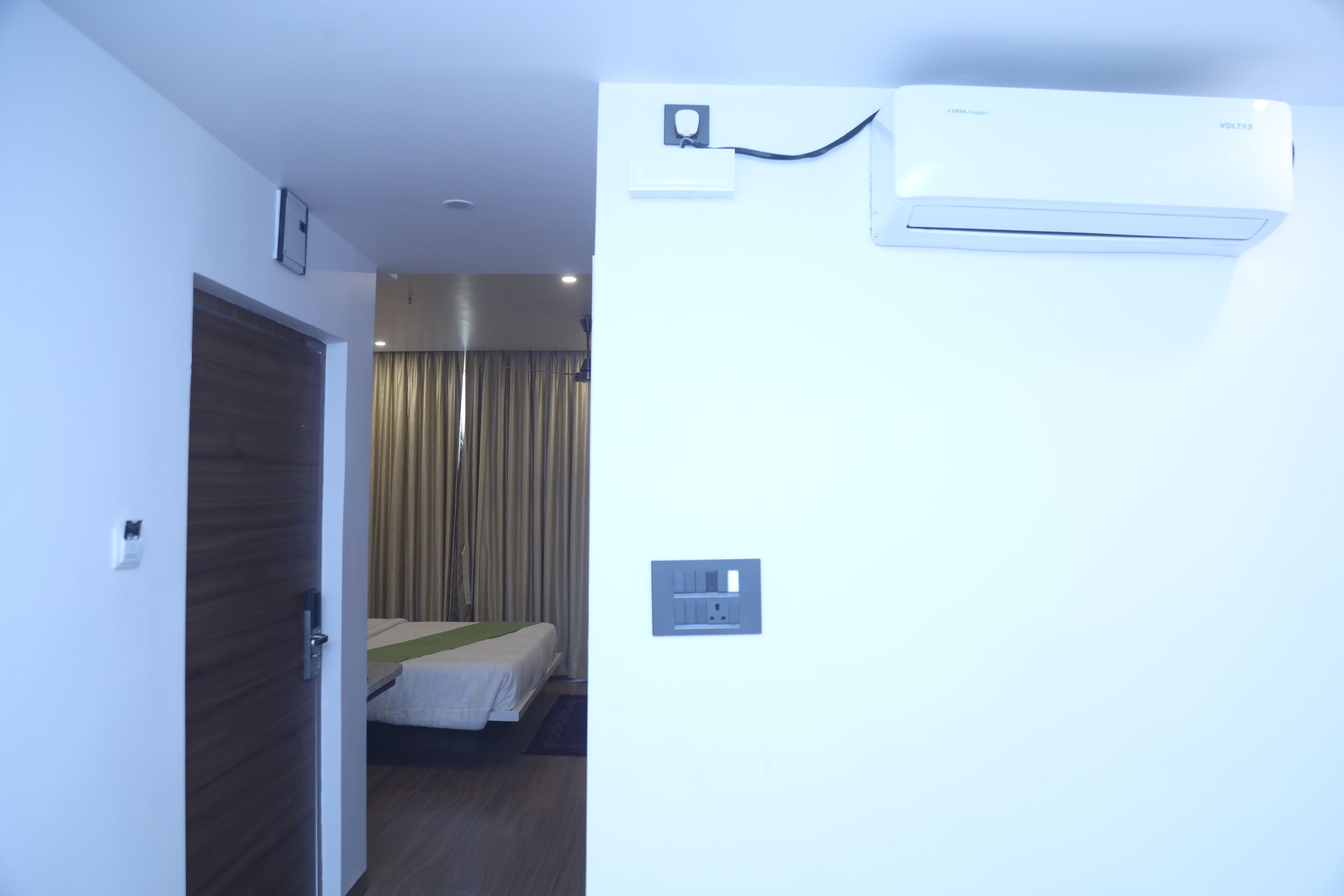 A hotel room with a bed, a door, an air conditioning unit, and a power outlet on the wall.