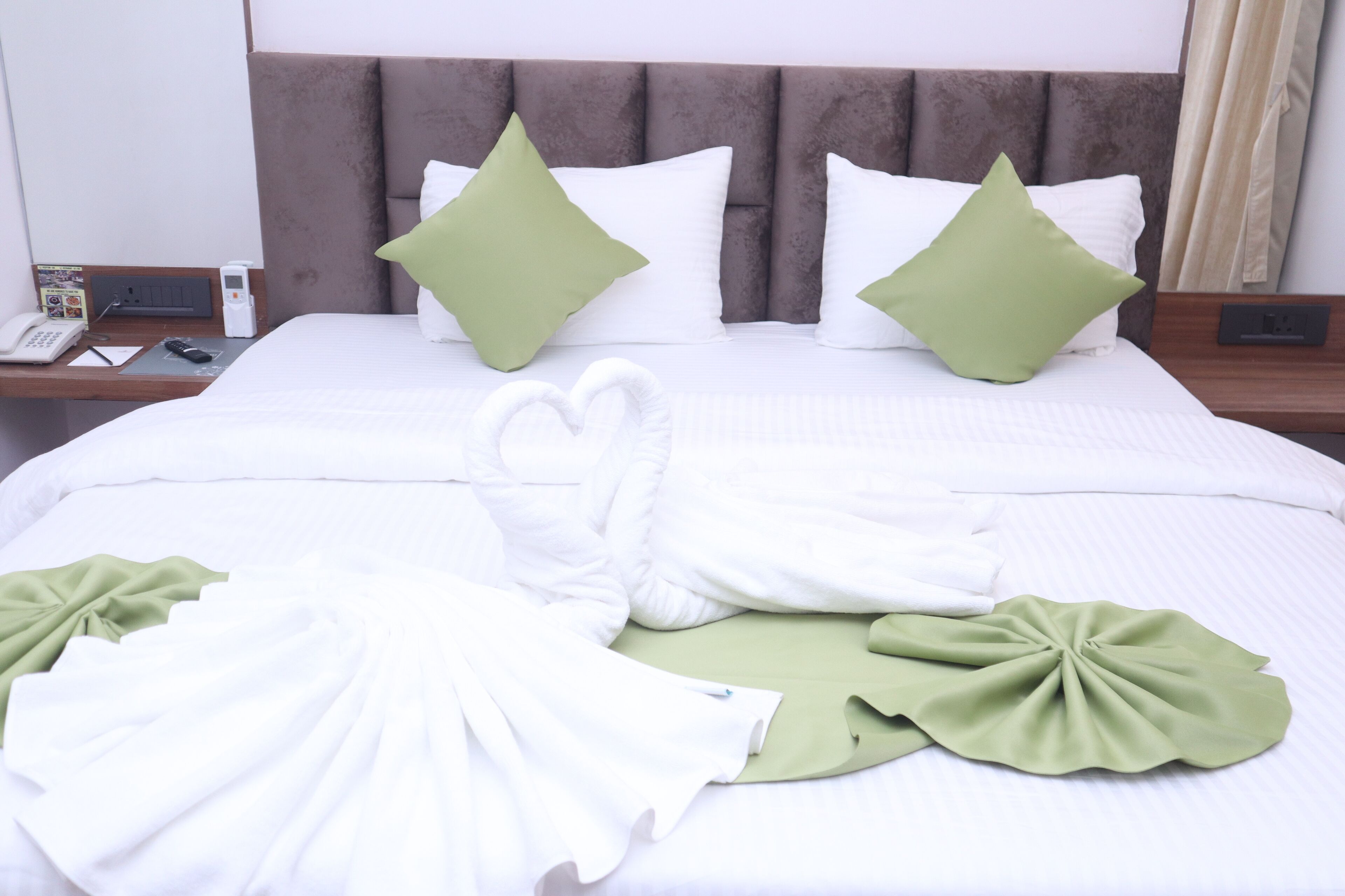 A neatly made bed with white and green linens, a heart-shaped towel arrangement, and a brown headboard.