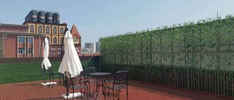 A rooftop deck with a wooden floor, metal chairs, and a table. There are umbrellas and a clock on the building in the background.