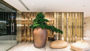 A modern lobby with a large bronze vase containing a green tree, two wooden poufs, and a metallic wall with vertical lines. - Full Season Hotel (Qingdao)