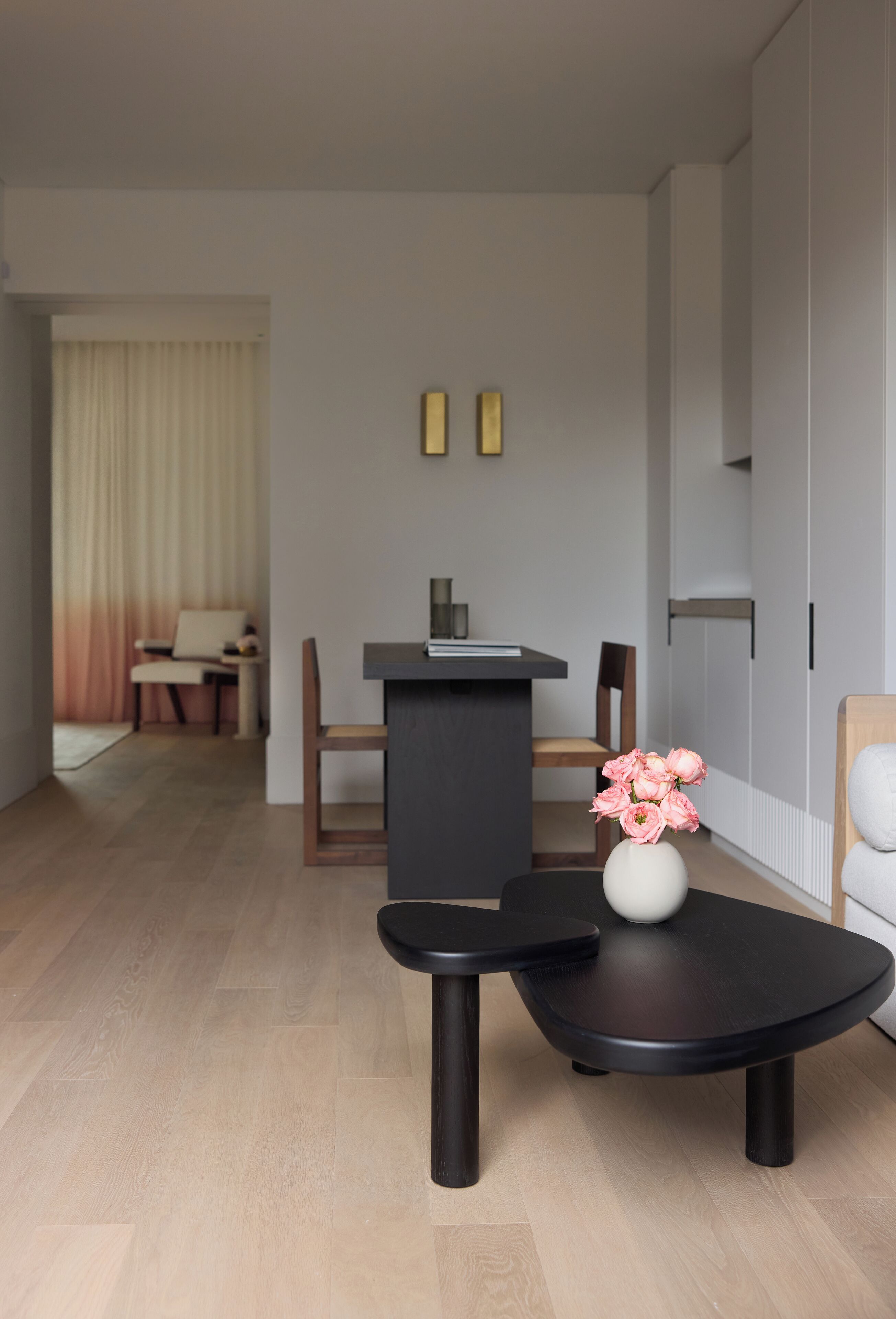 Ein modernes Wohnzimmer mit einem dunkelholzigen Couchtisch, einer weißen Vase mit rosa Blumen, einem schwarzen Beistelltisch und einem dunkelholzigen Sessel.