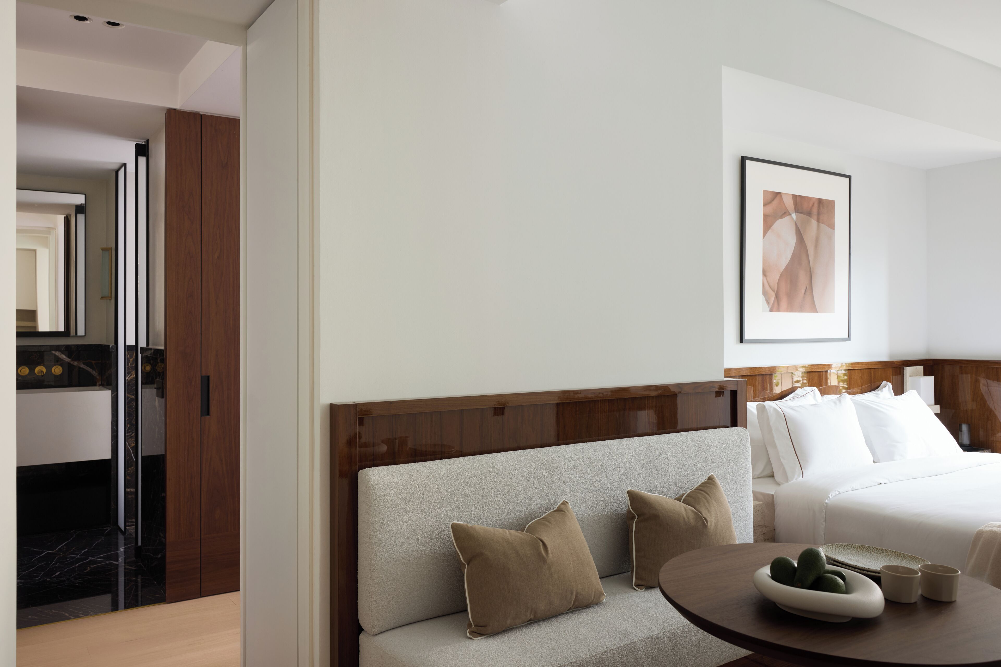 Une chambre d’hôtel avec un canapé, une table ronde sur laquelle se trouve un saladier de fruits, un lit avec une literie blanche et un tableau encadré au mur.