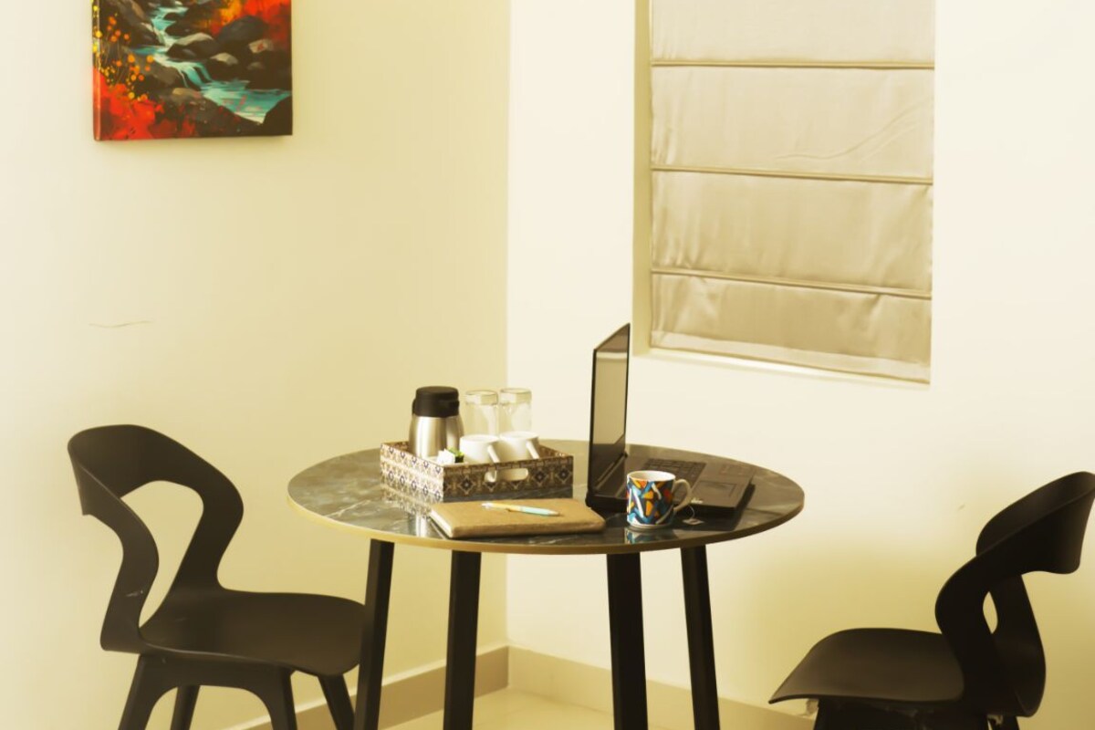 Uma área de refeições com uma mesa redonda, duas cadeiras pretas, um ventilador de teto, uma luminária pendente, um quadro e uma janela com persianas.