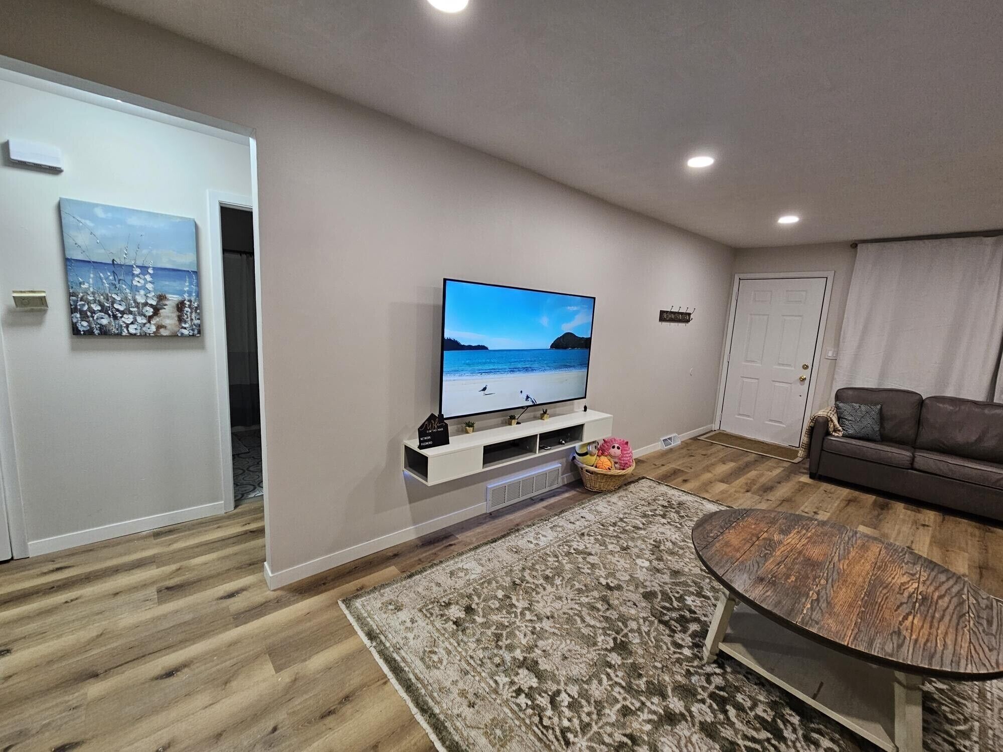 A modern living room with a flat-screen TV, wooden flooring, a patterned rug, and a brown leather sofa.