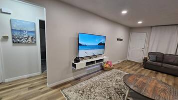 A modern living room with a flat-screen TV, wooden flooring, a patterned rug, and a brown leather sofa.