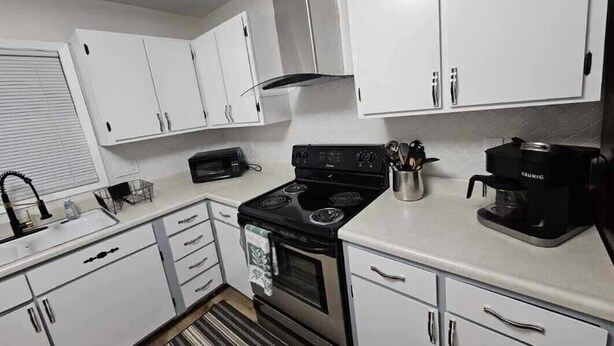 Une cuisine de taille réduite avec des meubles blancs, une cuisinière noire et une machine à café.