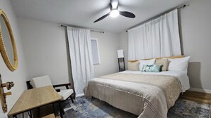 Un dormitorio con cama, escritorio, silla, ventilador de techo y ventana con cortinas.