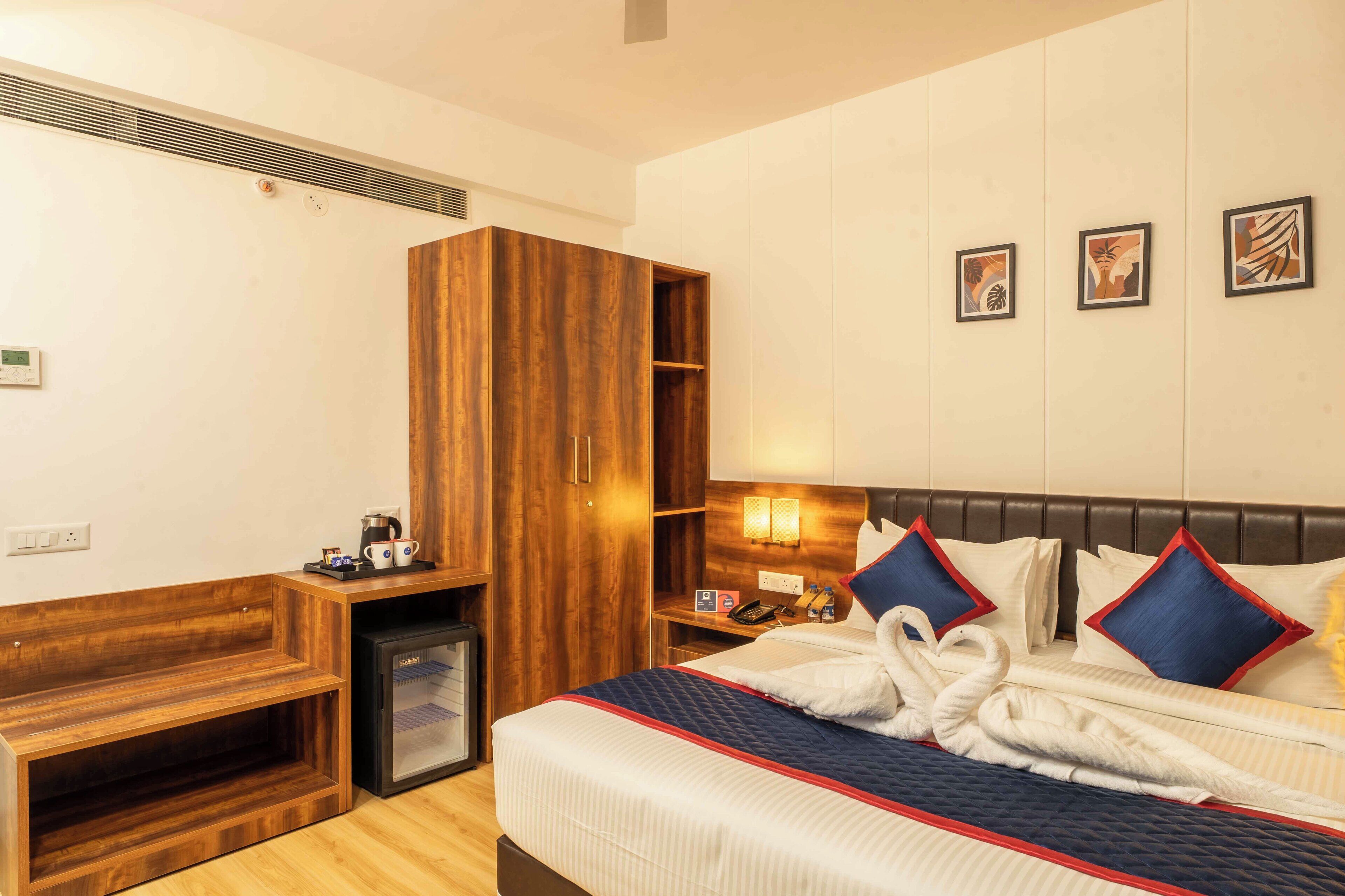 Une chambre d’hôtel comprenant un lit, une armoire en bois, un petit réfrigérateur, une table de chevet avec une machine à café et des œuvres d’art accrochées au mur.