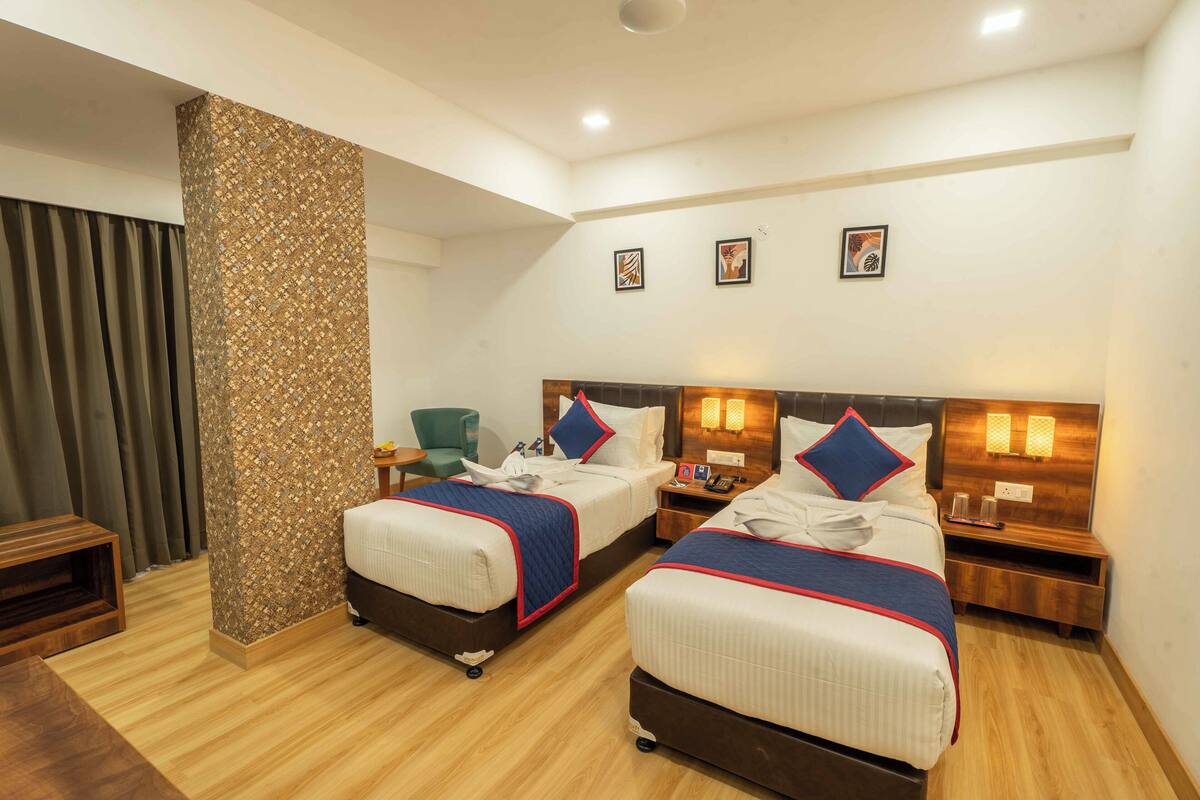 A hotel room with two beds, a wooden floor, a large pillar, and three wall-mounted lights.