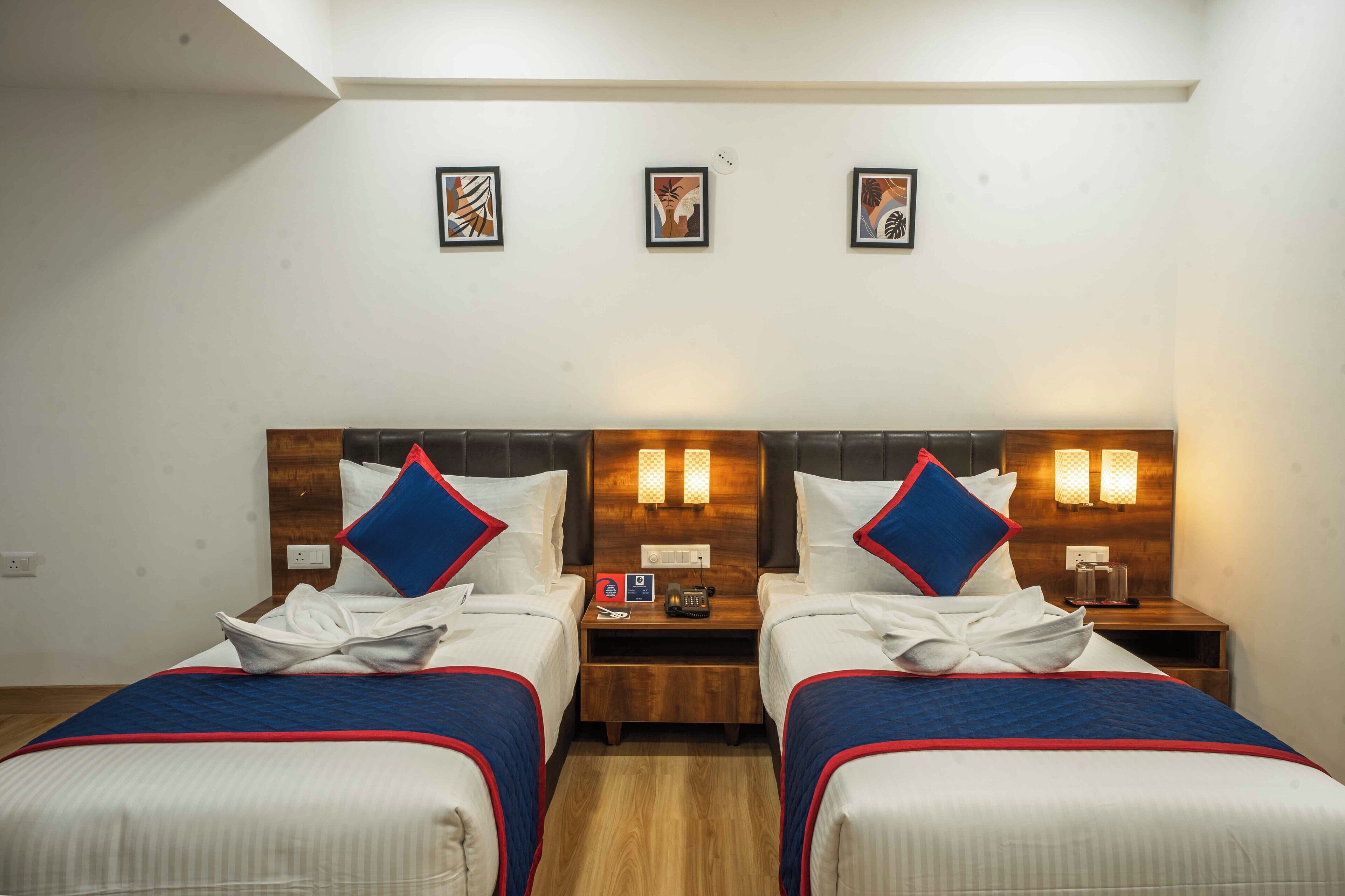 Habitación de hotel con dos camas, cada una con cabecero de madera, ropa de cama blanca y almohadas azules y rojas. Hay luces fijadas en la pared sobre las camas, una mesita de noche pequeña con un teléfono y una taza, y tres cuadros enmarcados en la pared.