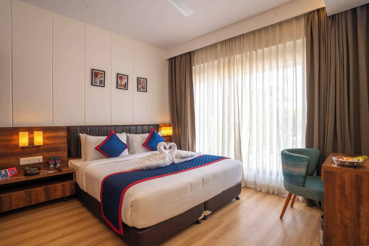 A hotel room with a large bed, a bedside table with a lamp, a chair, and a window with curtains.