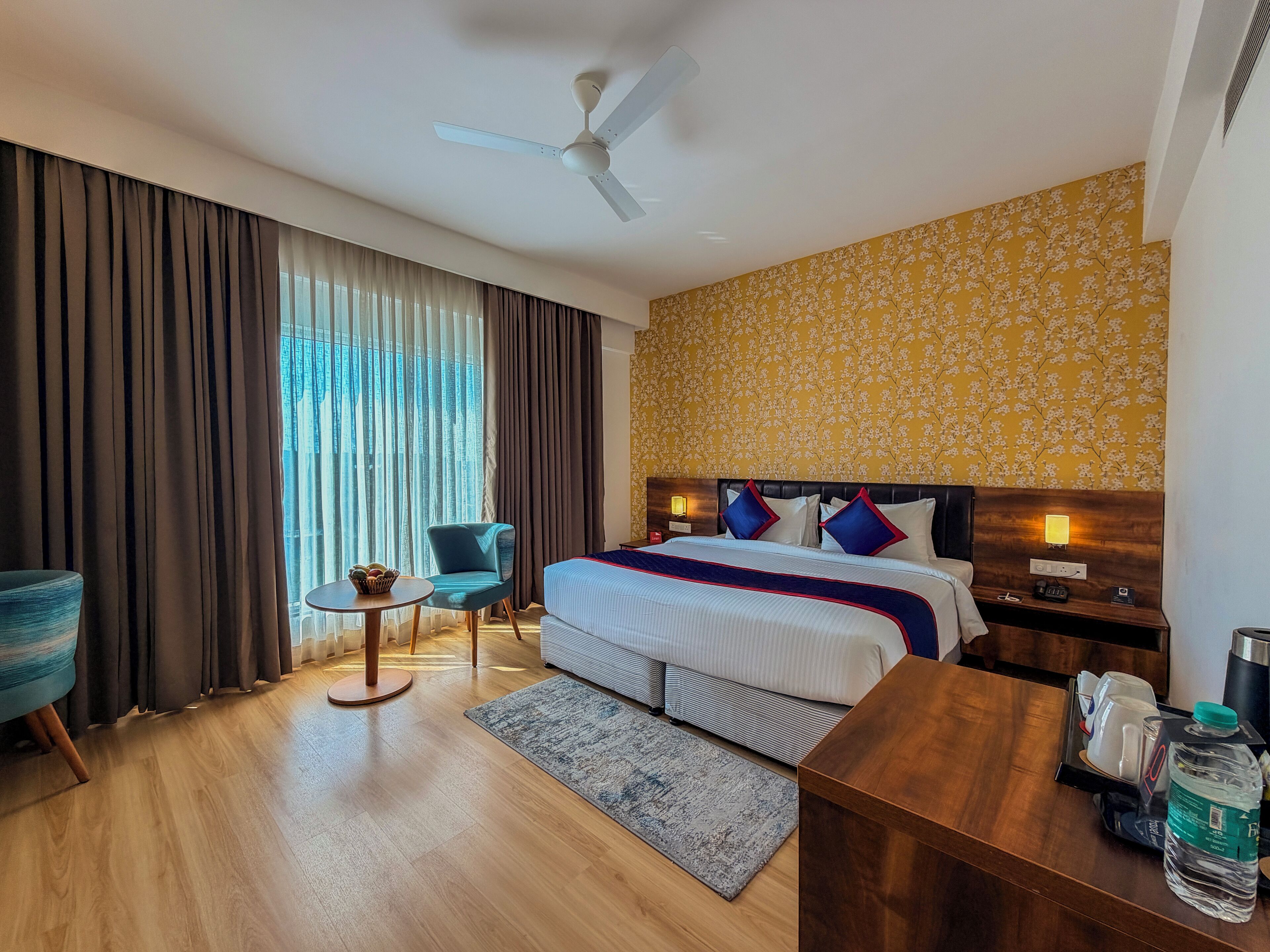 A hotel room with a bed, a desk, a chair, a small table, and a ceiling fan.