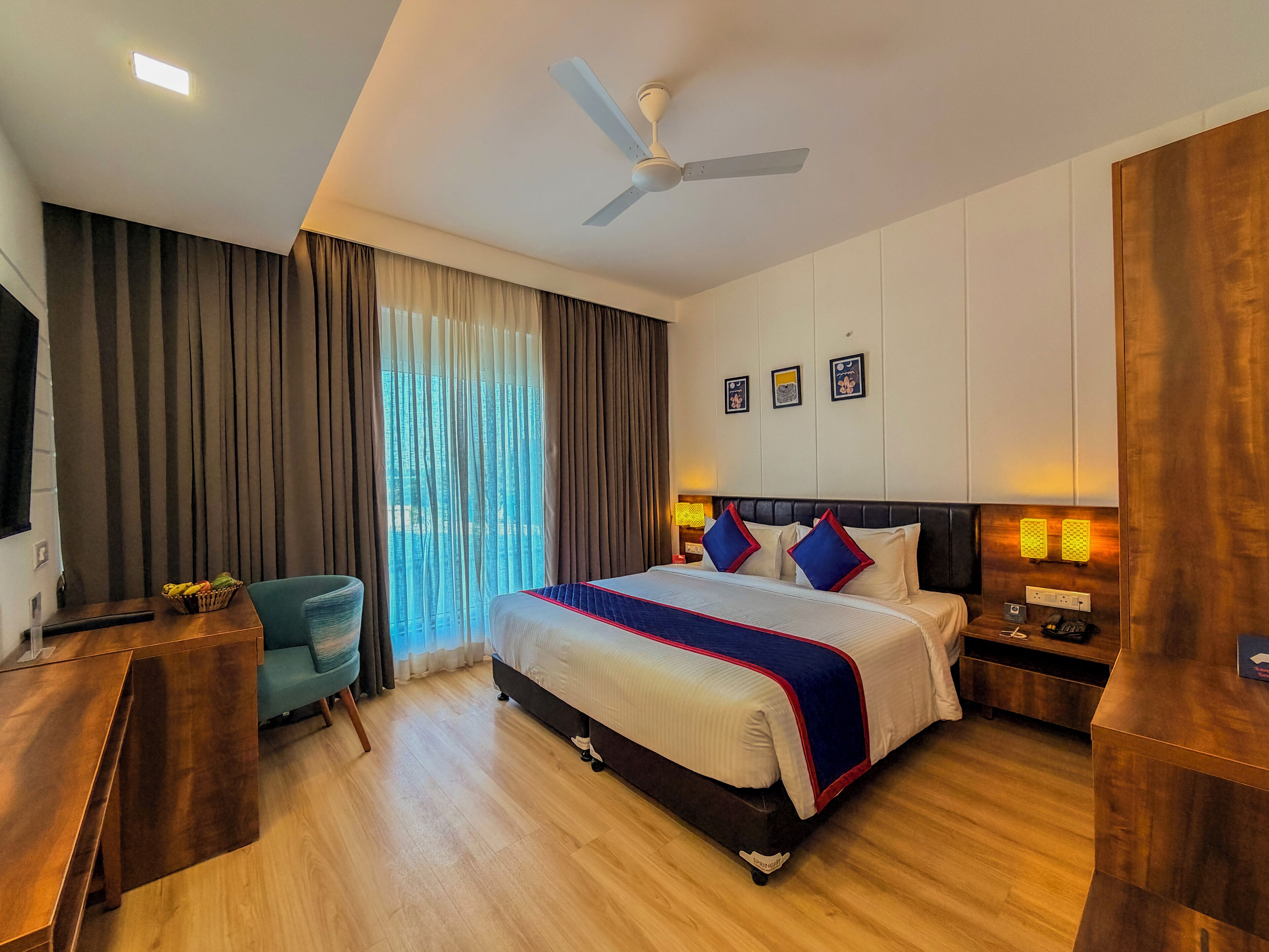 A hotel room with a bed, a desk, a chair, a TV, a ceiling fan, and a window with curtains.