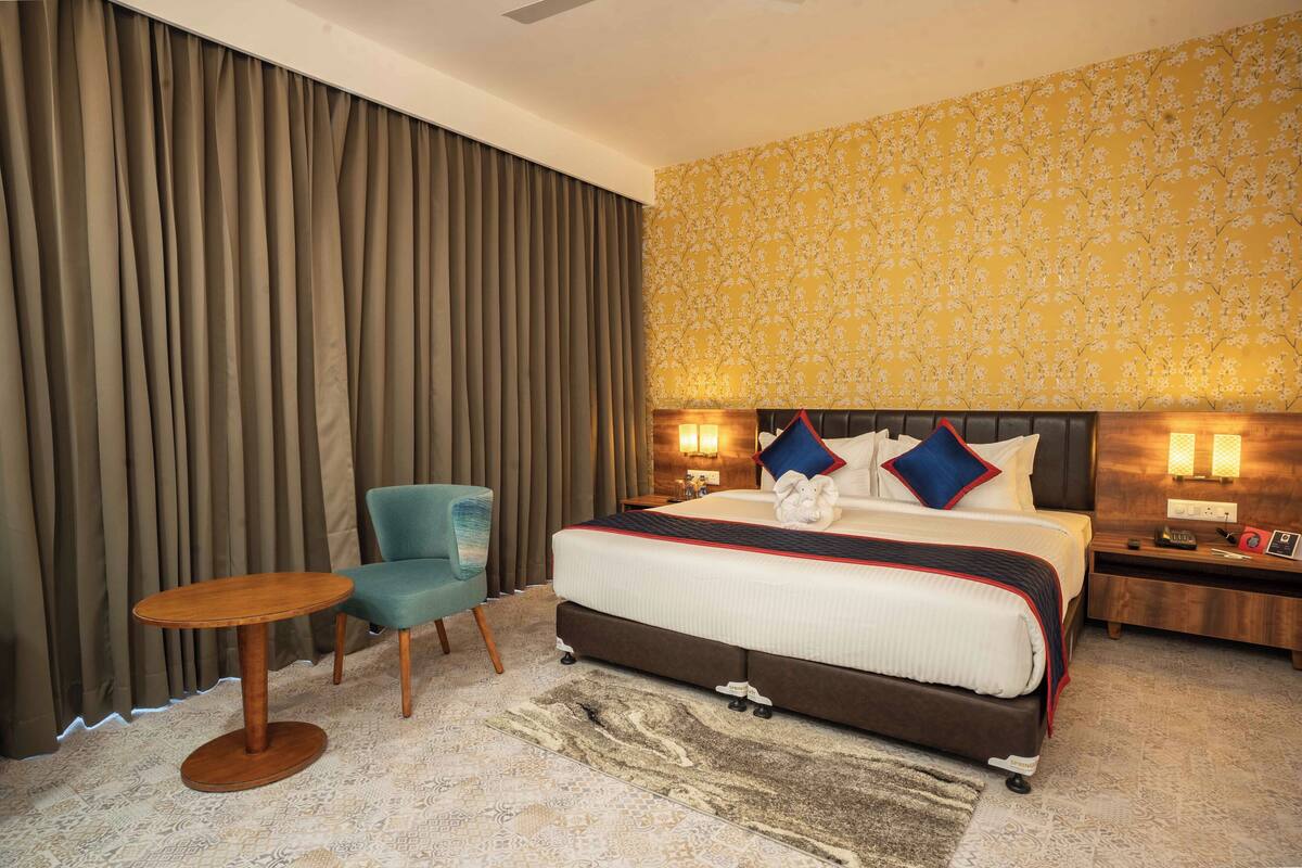 A hotel room with a large bed, a small round table, a blue chair, and a nightstand with a lamp.