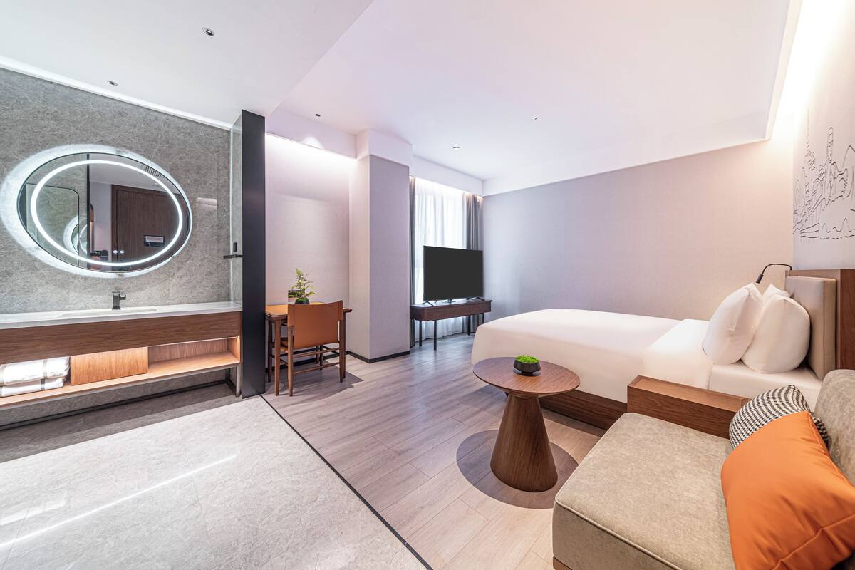 A modern hotel room with a large round mirror, a sofa, a small table, a TV, and a desk with a chair.
