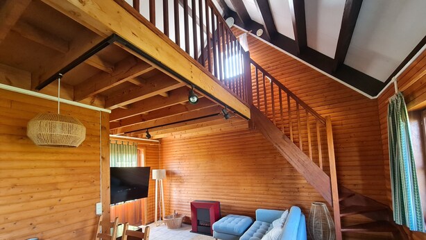 Interior de una cabaña de madera con escalera, sofá, televisor y comedor.