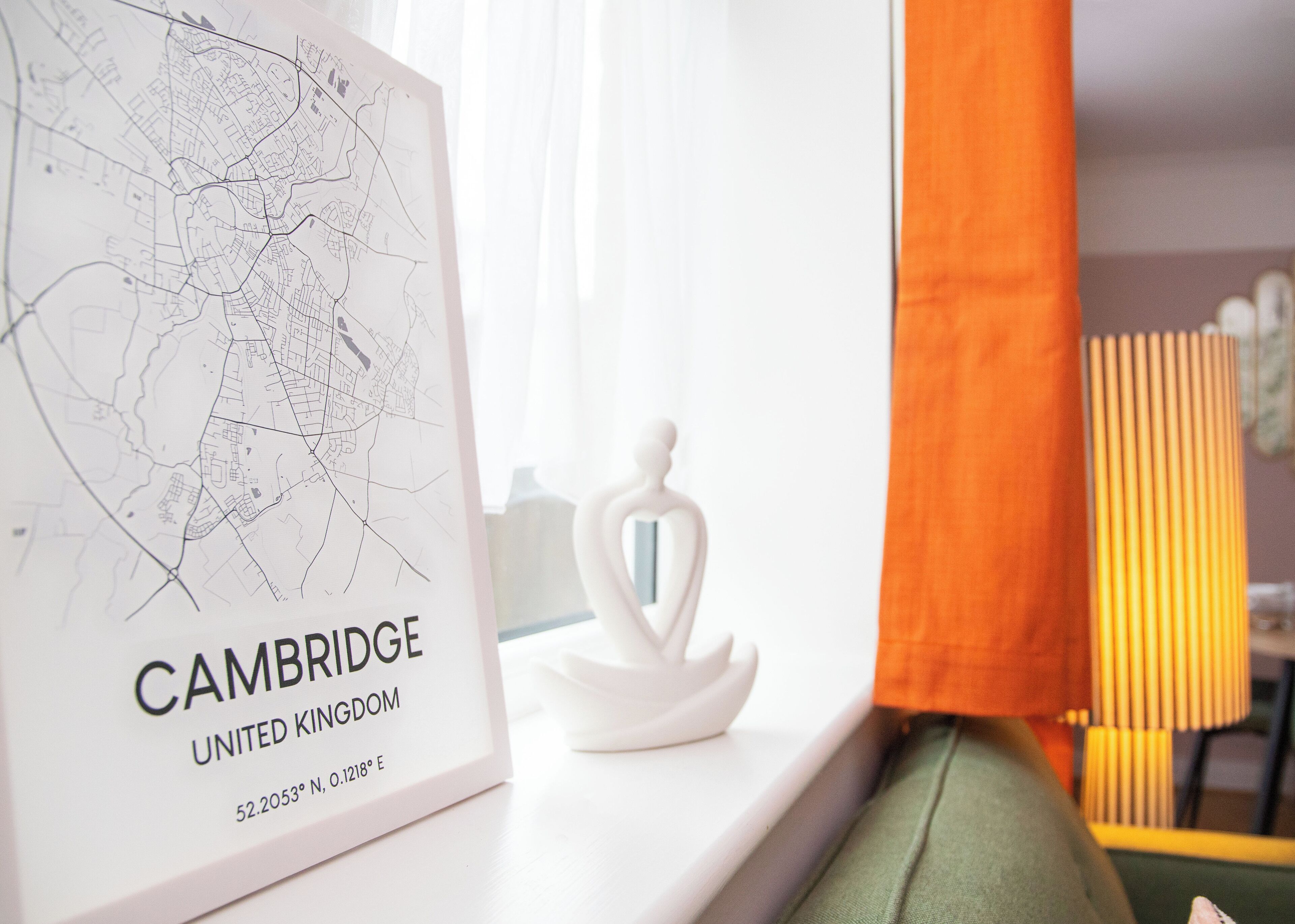 Een ingelijste kaart van Cambridge, Verenigd Koninkrijk, met de coördinaten 52.2053° N, 0.1218° E, op een wit oppervlak, naast een witte decoratie en een raam met oranje gordijnen.