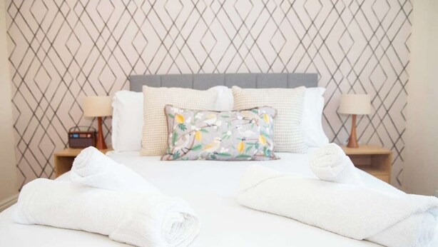 Una cama bien hecha con sábanas blancas y un cojín estampado con motivos florales, flanqueada por dos lámparas de noche sobre mesitas de madera.