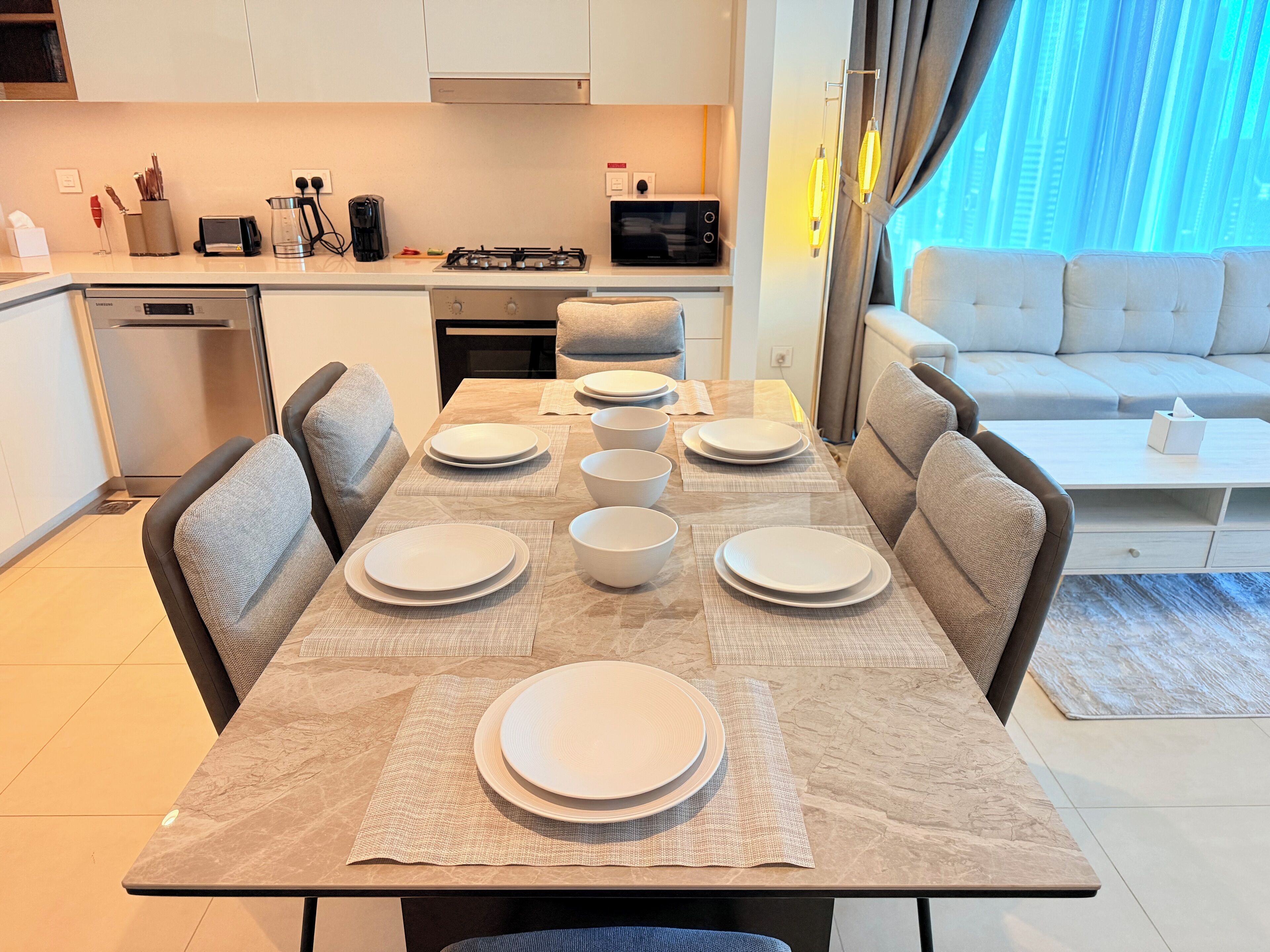 Une cuisine moderne avec un coin repas, comprenant une table dressée avec des assiettes et des bols, un canapé et une vue sur la ville grâce à de grandes fenêtres.