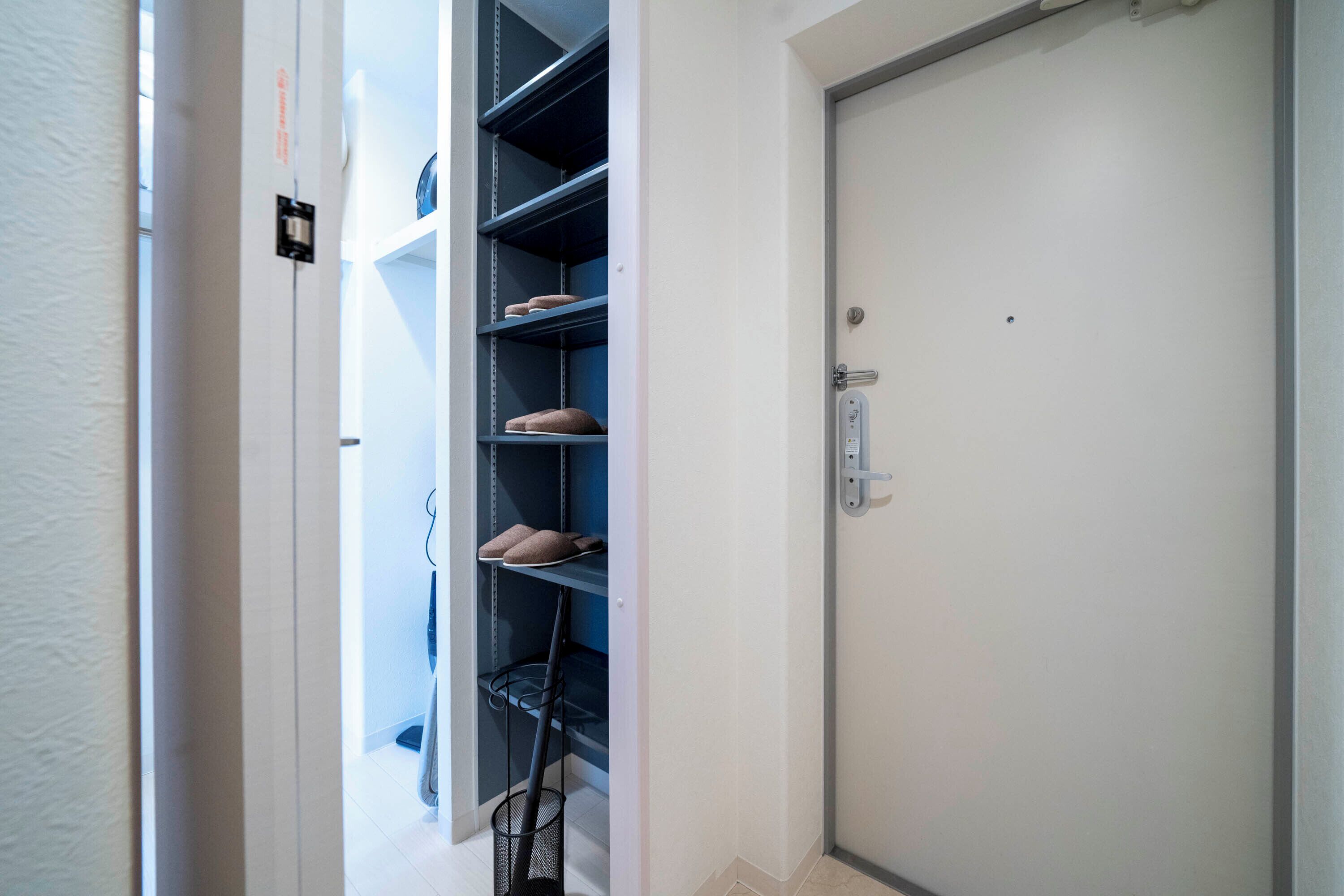 Un couloir avec une porte fermée, un porte-chaussures rempli de chaussures et un balai appuyé contre le mur.