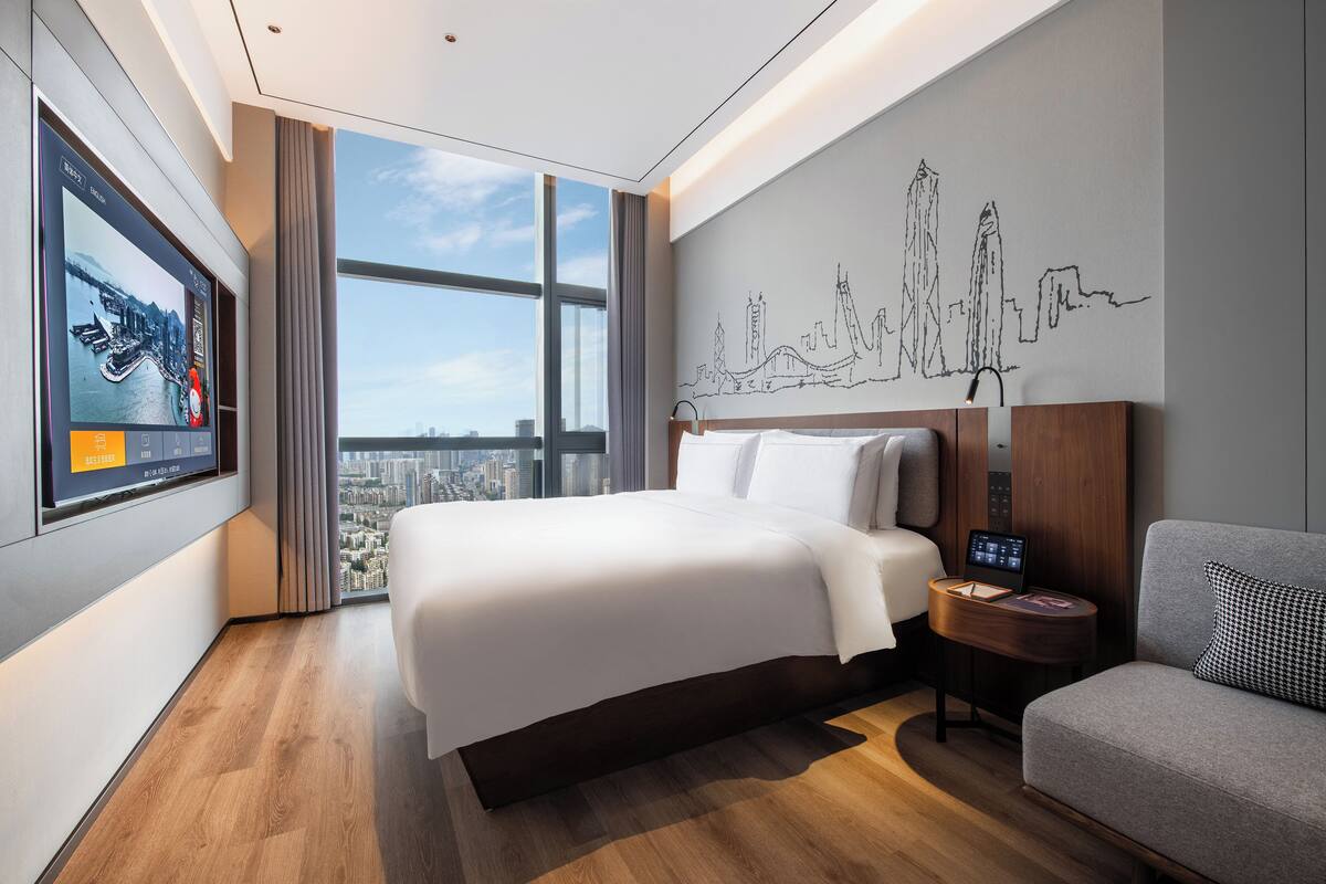 A modern hotel room with a large bed, a city view, a wall art of a city skyline, and a small table with a laptop.
