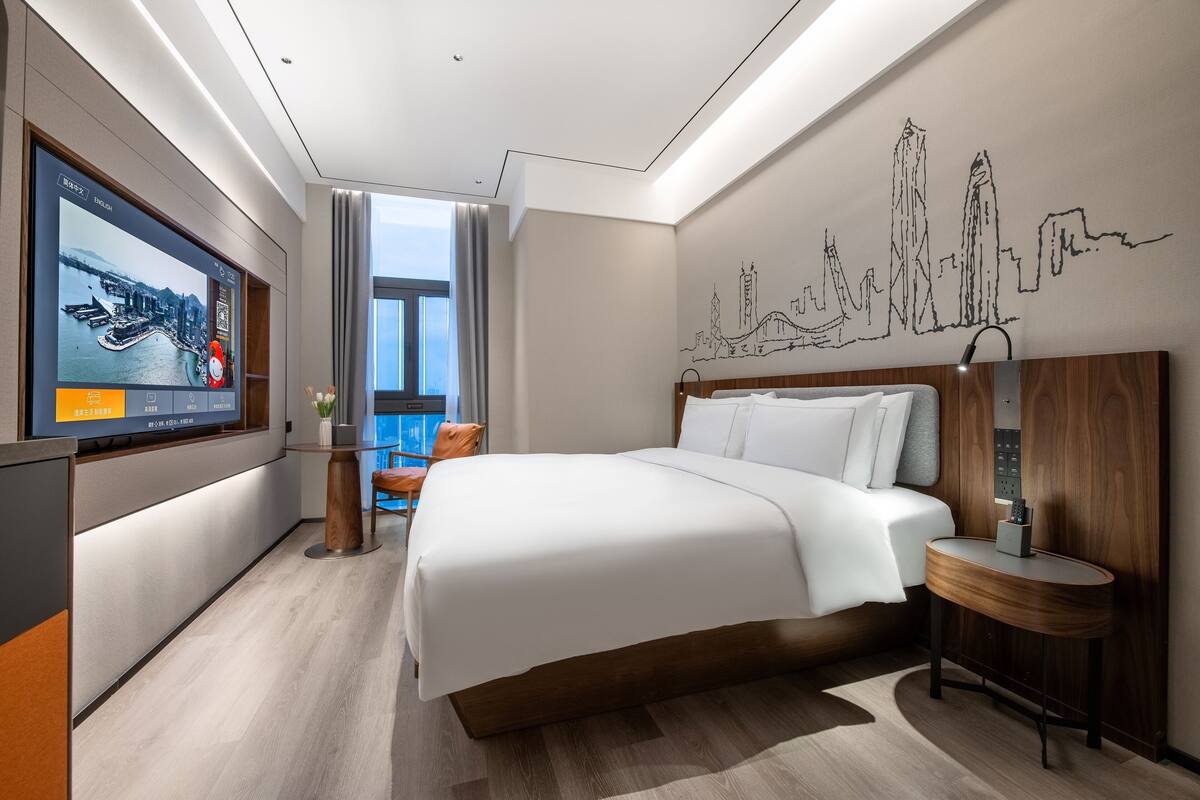 A modern hotel room with a large bed, a TV on the wall, a city skyline mural, and a view of the outdoors.