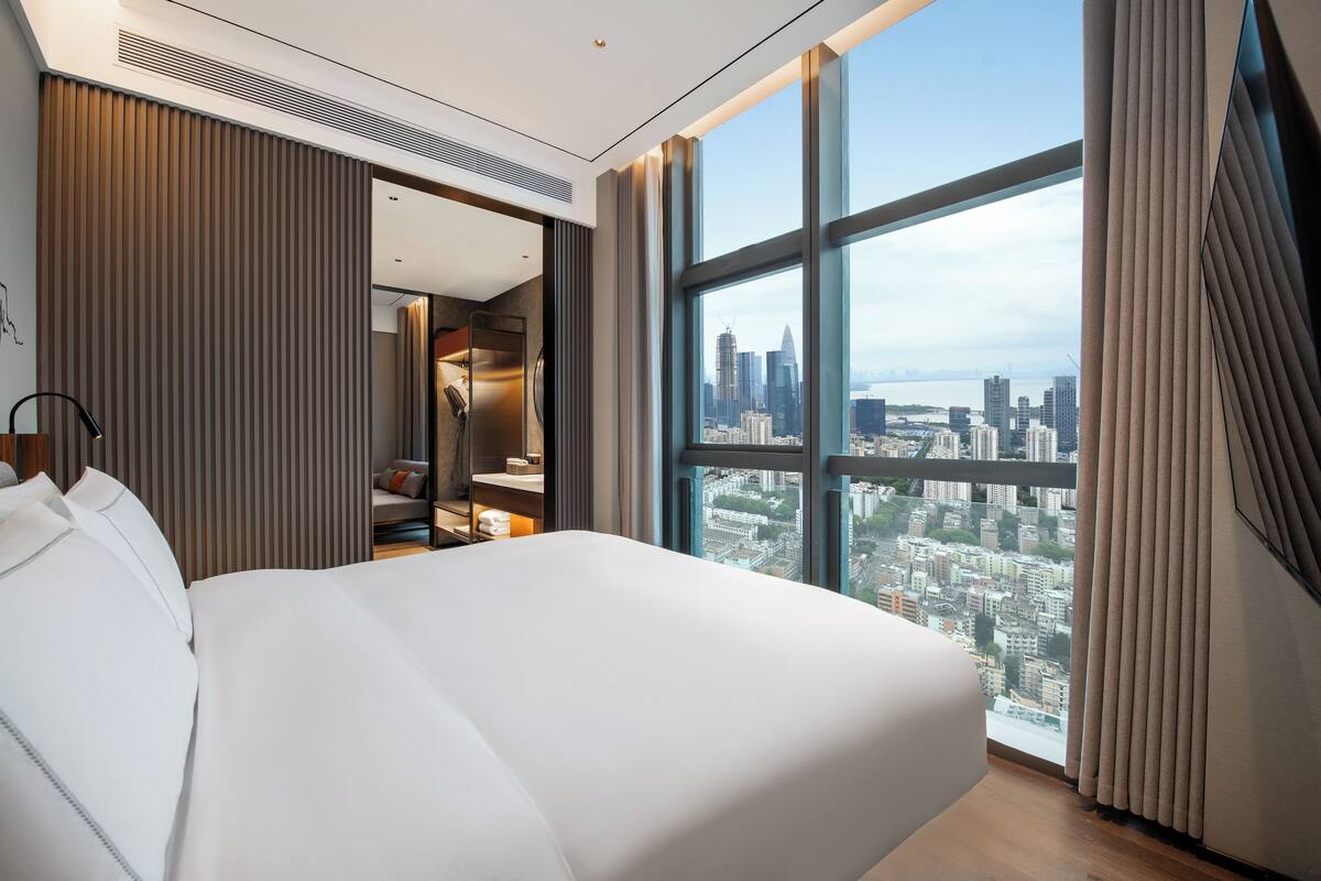 A modern hotel room with a large bed, a city view, and a walk-in closet.