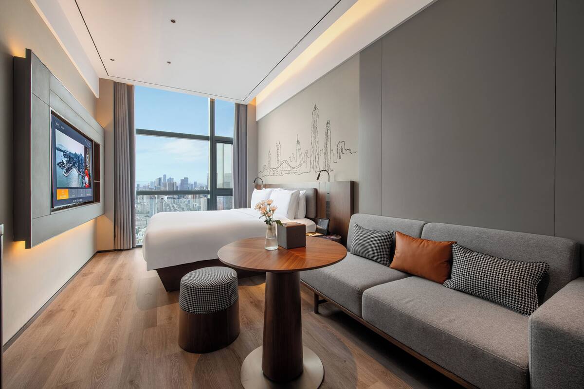 A modern hotel room with a bed, a gray sofa, a round wooden table, and a city view through large windows.