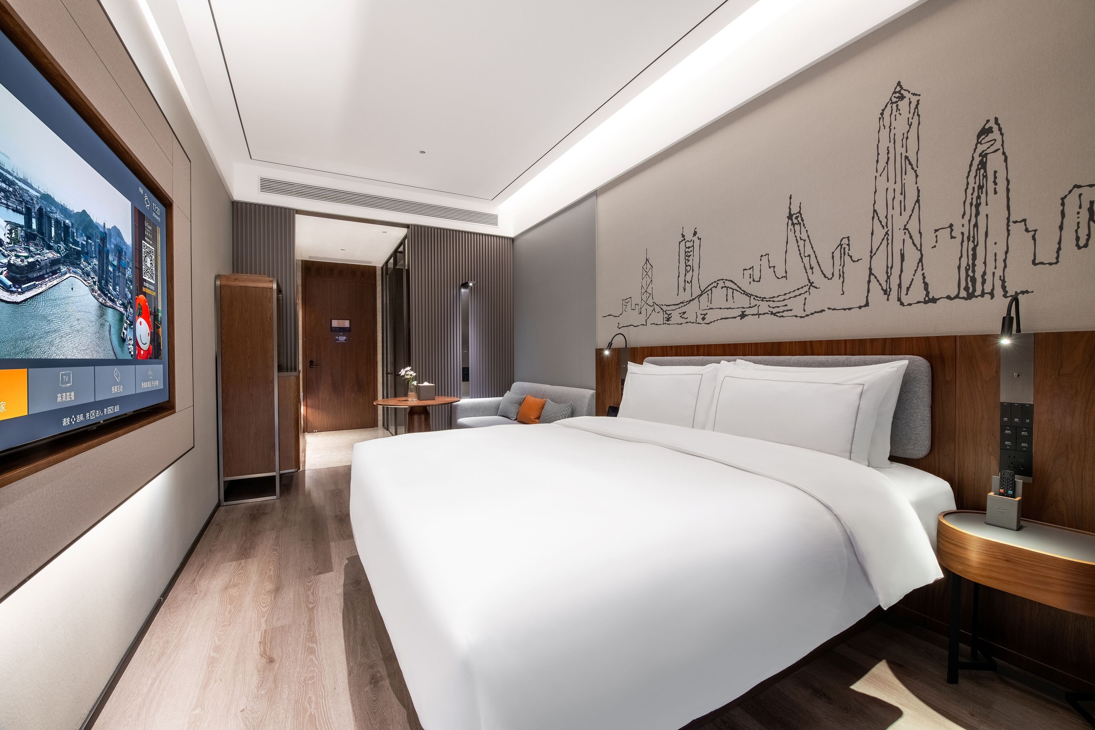 A modern hotel room with a large bed, a flat-screen TV, a cityscape wall art, and a wooden headboard.