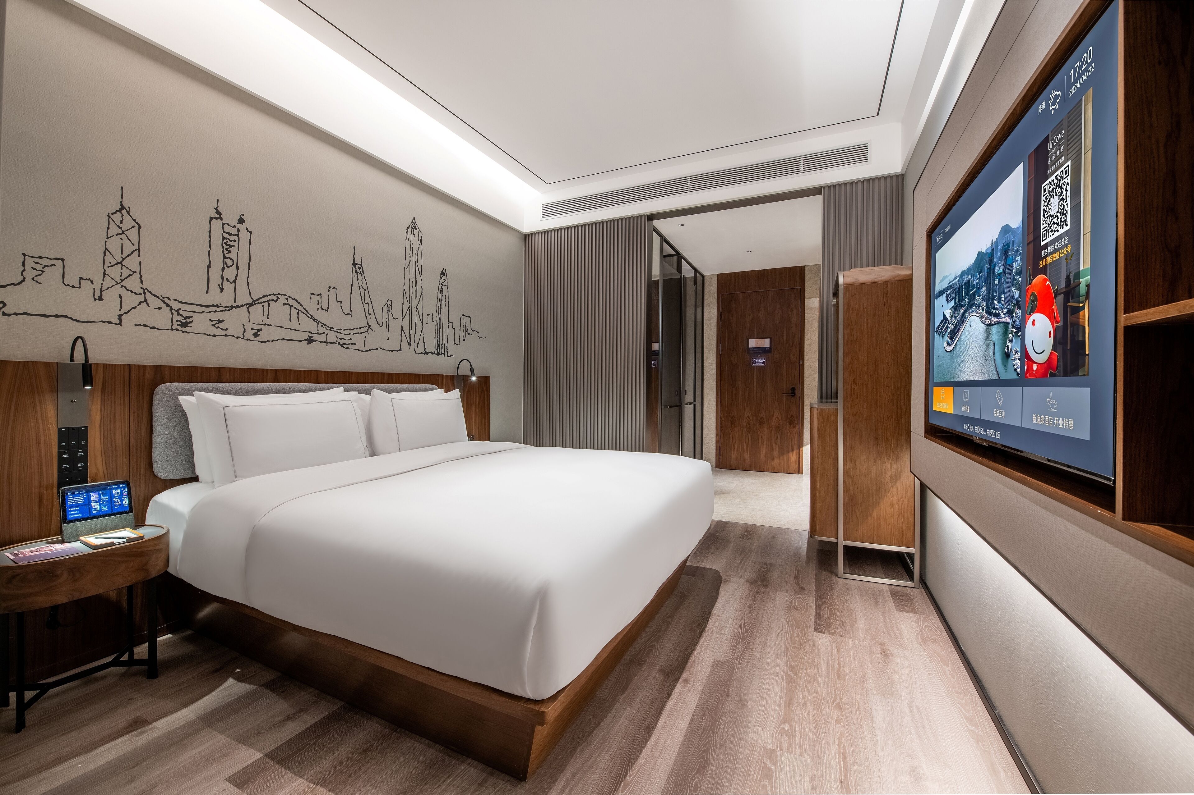 Une chambre d’hôtel moderne dotée d’un grand lit, d’un bureau avec un ordinateur portable, d’un téléviseur fixé au mur et d’une fresque représentant un paysage urbain sur le mur.