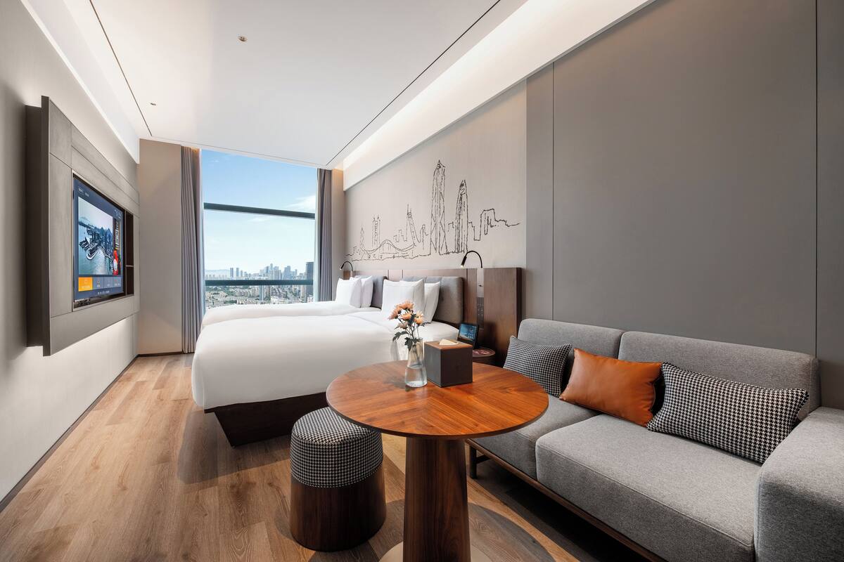 A modern hotel room with a large bed, a gray sofa, a round wooden table, and a city skyline view.