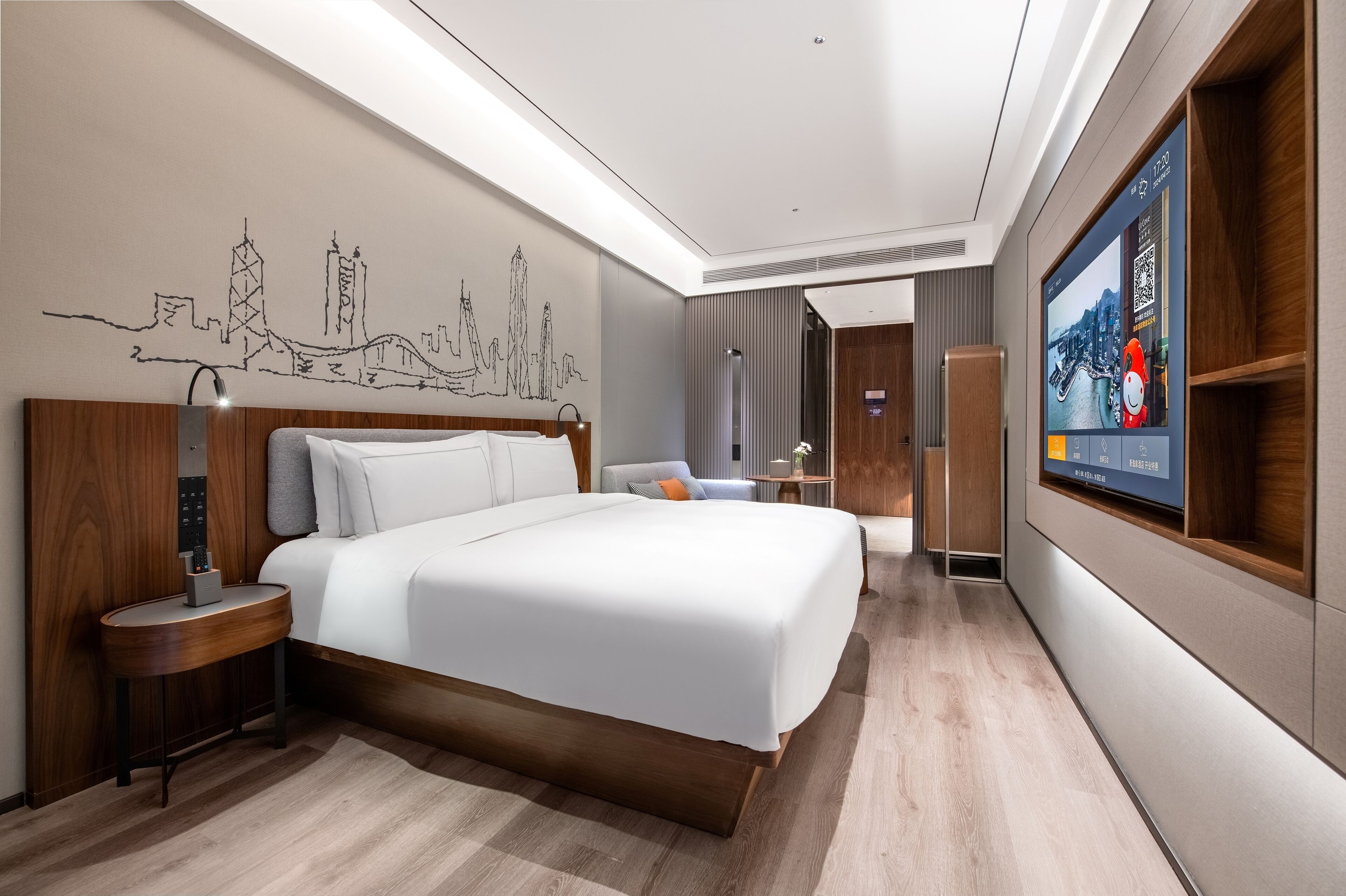 A modern hotel room with a large bed, a bedside table with a lamp, a wall-mounted TV, and a cityscape artwork.