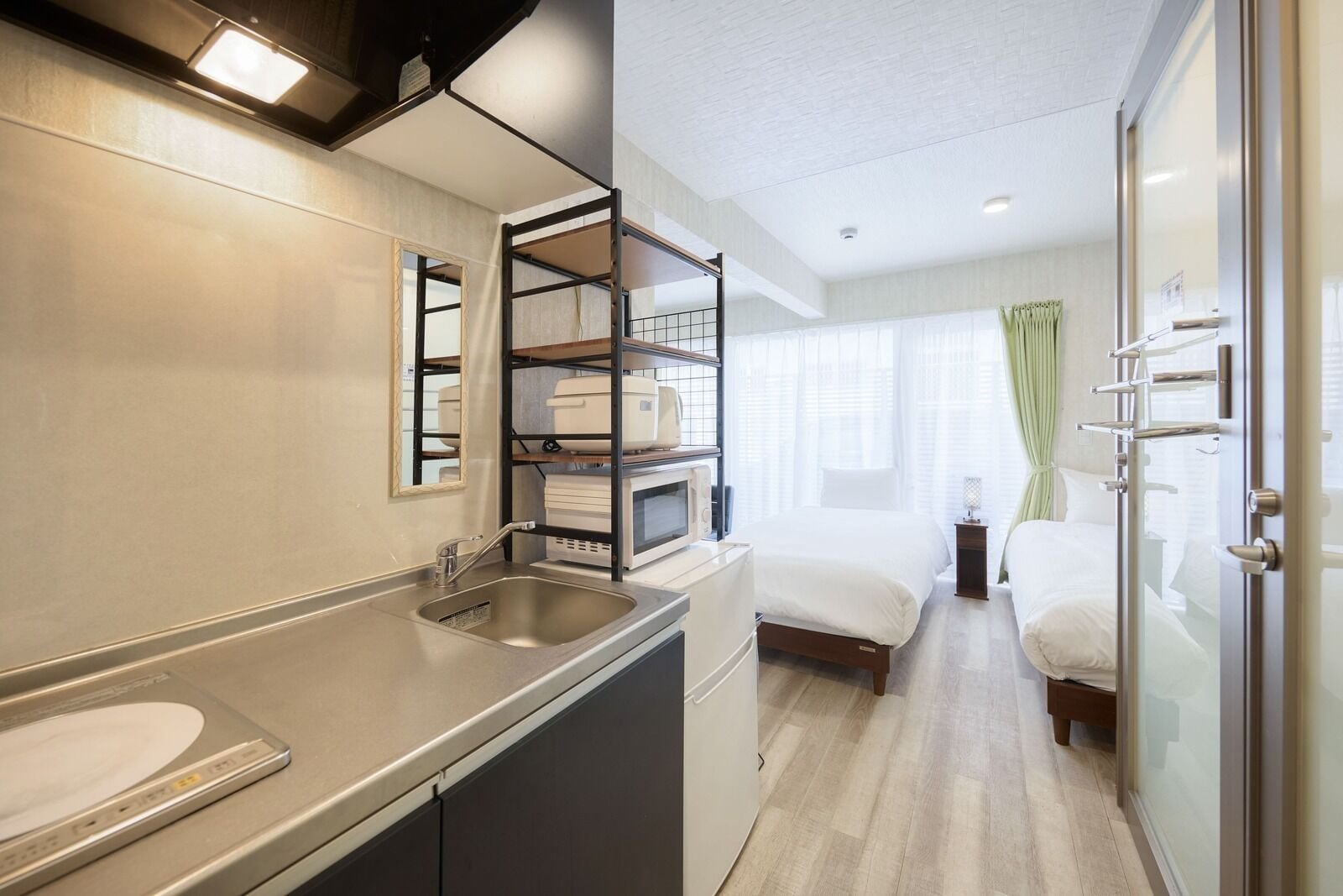 A compact hotel room with a kitchenette, a bed, a glass shower enclosure, and a view of the city.