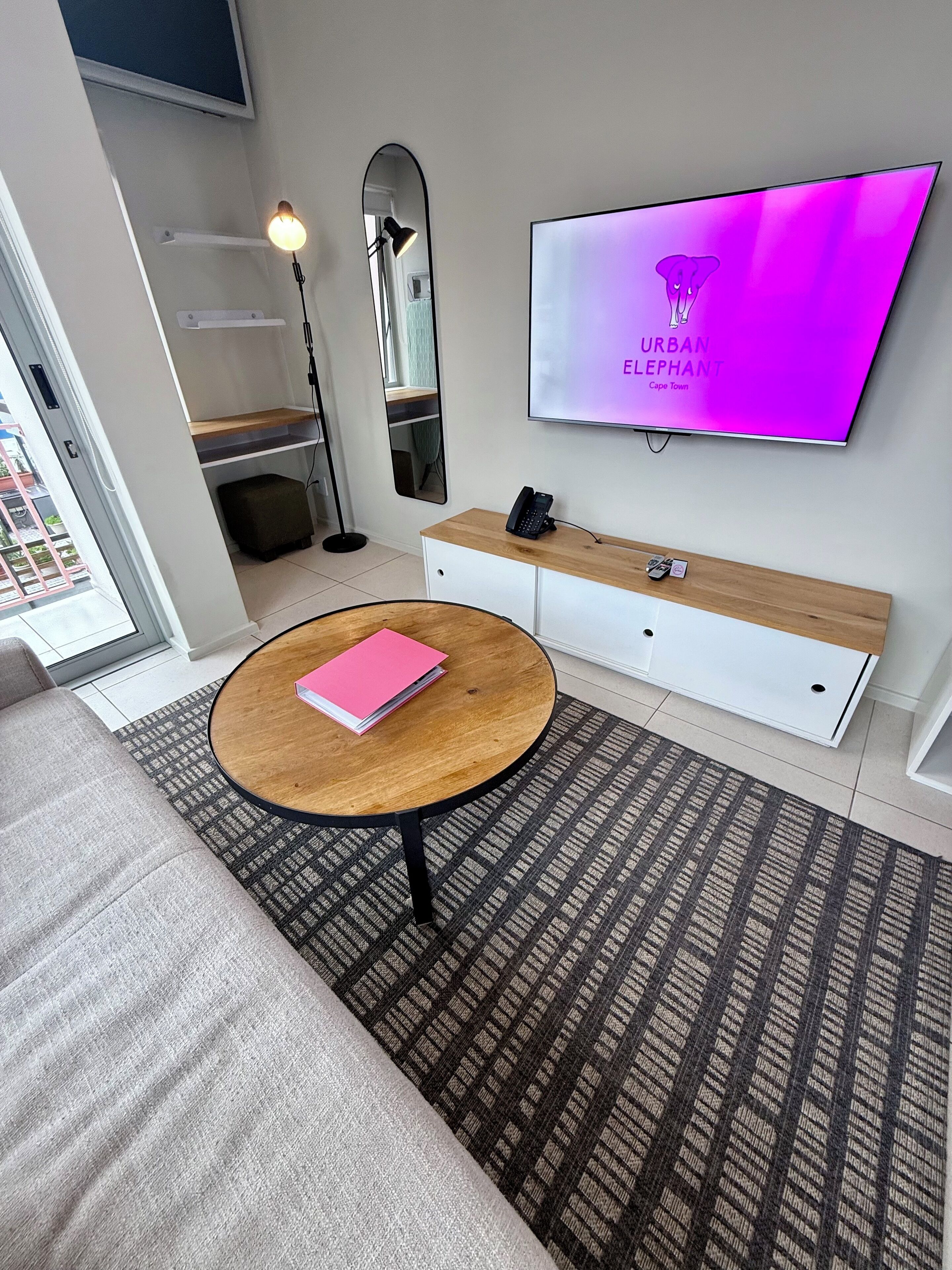 A modern living room with a round wooden coffee table, a grey sofa, a patterned rug, a white TV stand with a mounted TV displaying "URBAN ELEPHANT Cape Town," and a mirror reflecting the room.