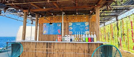 A beachside bar with bamboo walls, a menu board, and two turquoise chairs.
