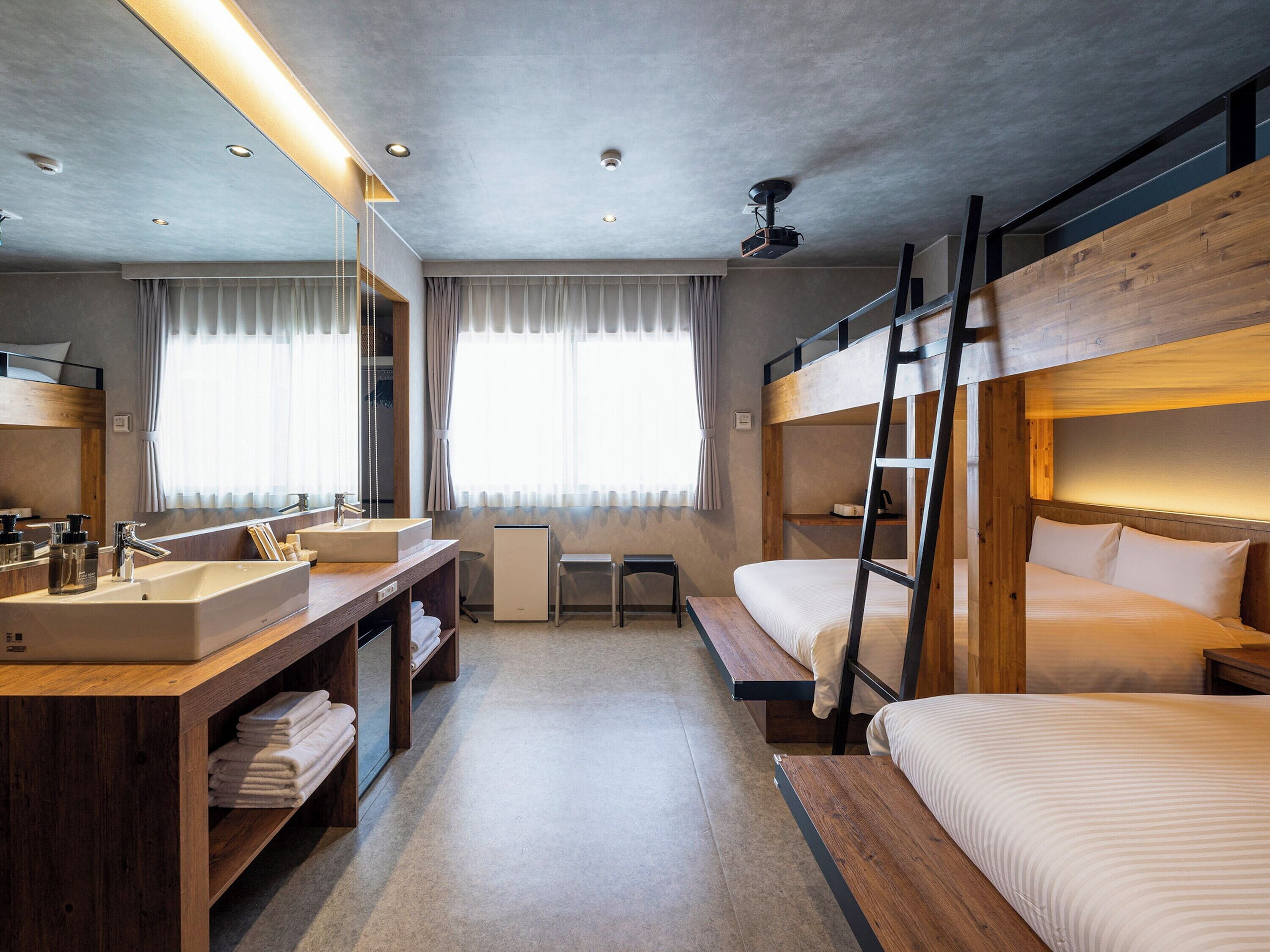 A modern hotel room with bunk beds, a large mirror, a wooden vanity with a sink, and a ceiling projector.
