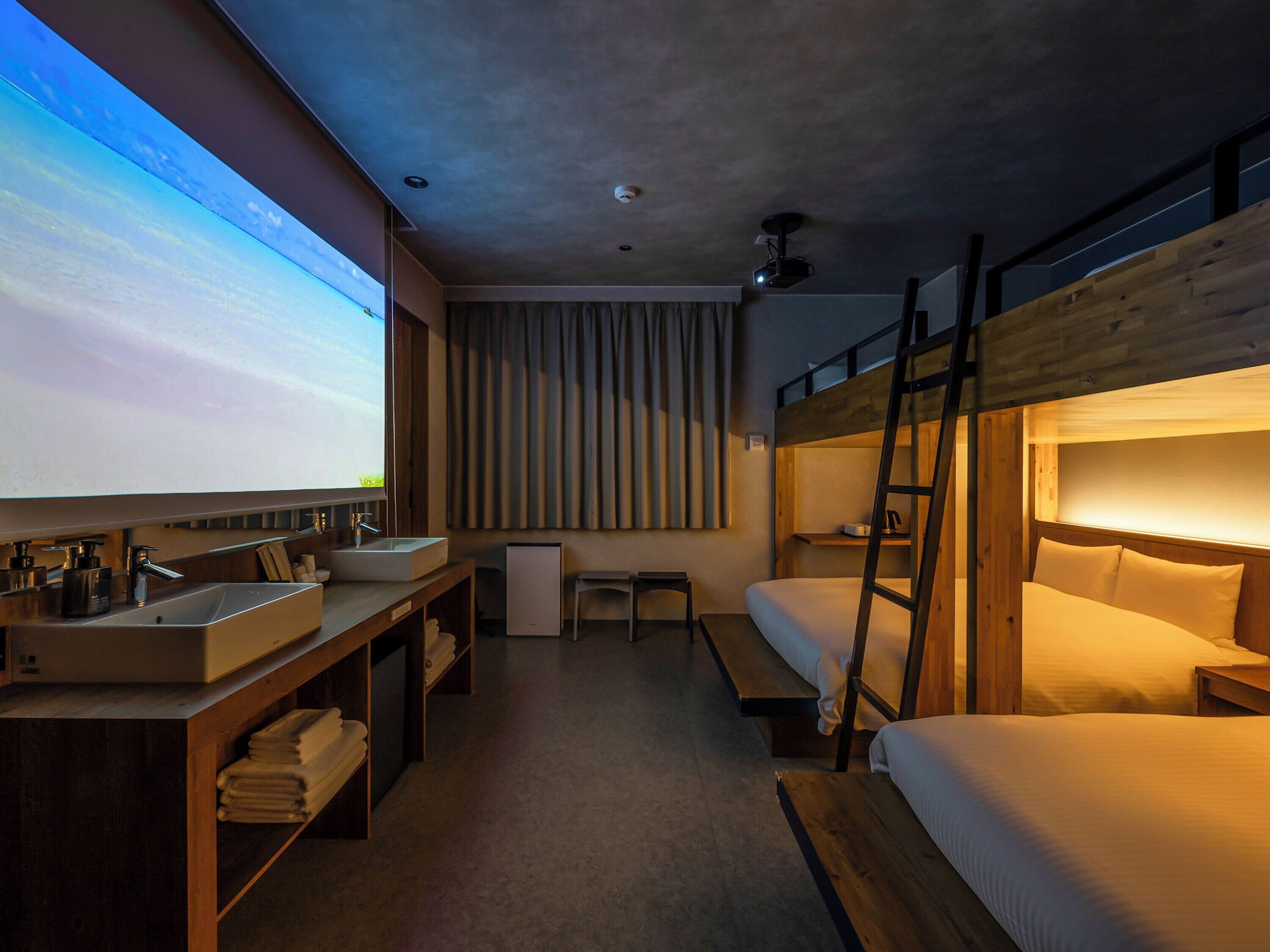 A modern hotel room with bunk beds, a large TV, a wooden desk with a sink, and a small table.