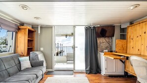 A compact RV interior with a gray sofa, a small kitchenette, and a view of the outdoors through a sliding door. - 62' Yacht M/v Possibilities On Wide Private Canal 3 Bedroom Houseboat (North Fort Myers)