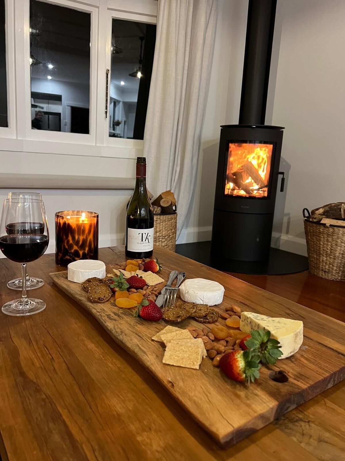 A cozy room with a wooden table set for a meal, a bottle of wine, and a candle. A wood-burning stove is visible in the background.