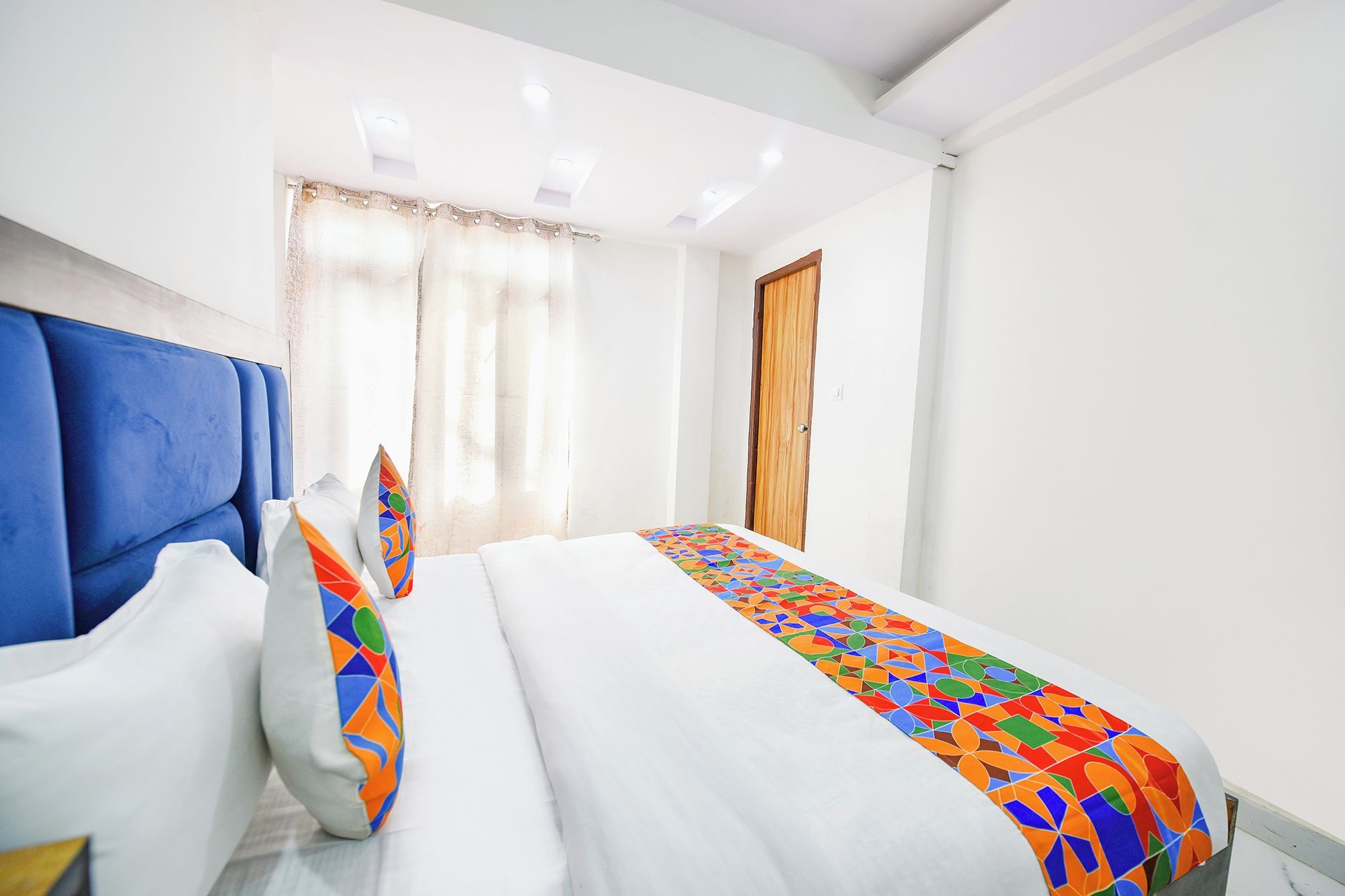 Habitación de hotel con una cama, dos almohadas, un cabecero azul, una puerta de madera y una ventana con cortinas.