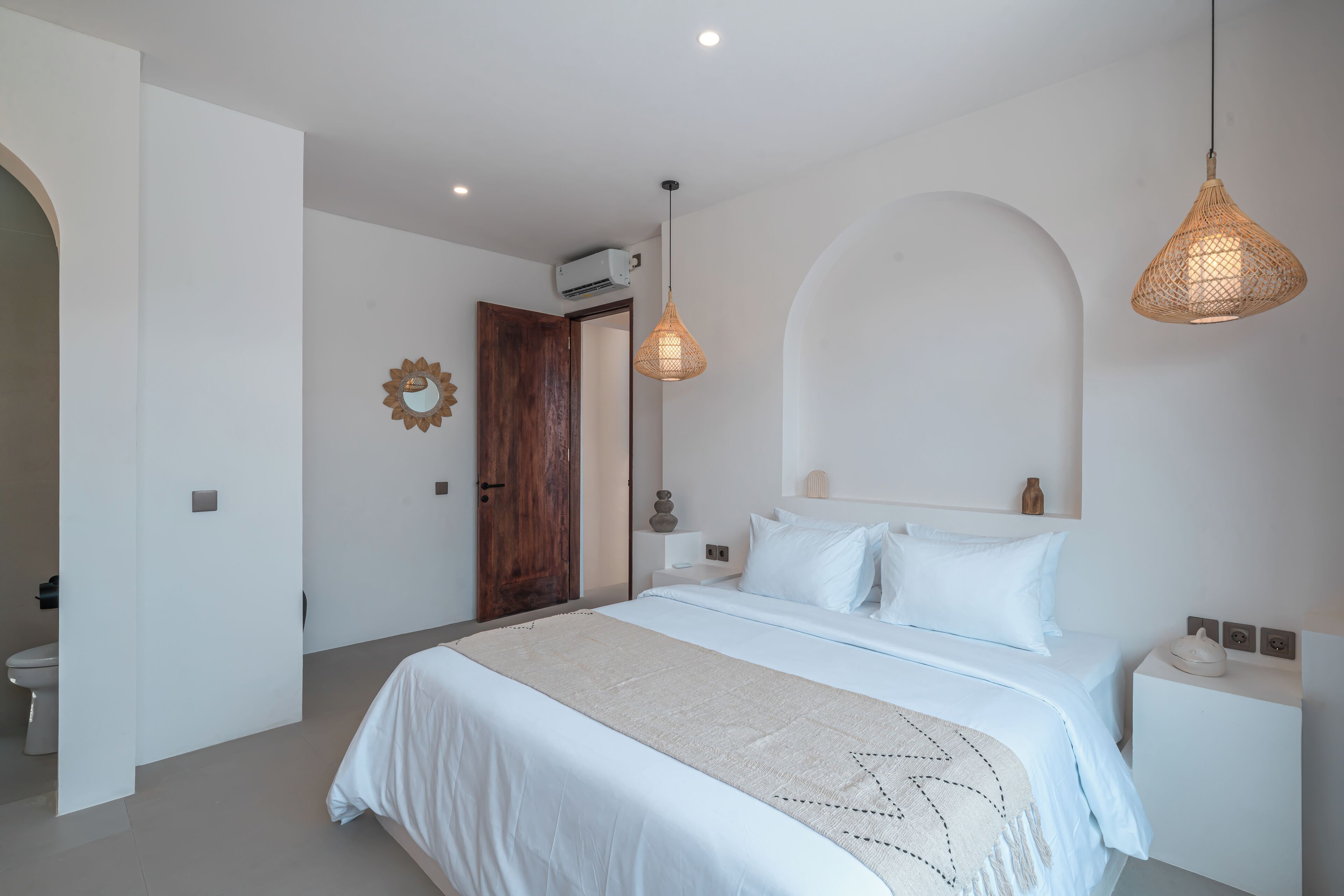 A bedroom with a bed, bedside tables, a hanging light fixture, and a door leading to another room.