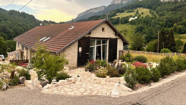 Uma casa de pedra com telhado inclinado, um pequeno alpendre e um jardim com várias plantas e flores.