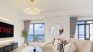 A modern living room with a large flat-screen TV displaying "NETFLIX", a sofa with patterned cushions, a round mirror, and a view of the city through large windows. - LUX  The Dubai Eye Sea View Suite (Dubai)