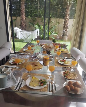 Une table de petit-déjeuner dressée avec différents plats et boissons, donnant sur un jardin agrémenté de palmiers et recouverte d’une nappe blanche.