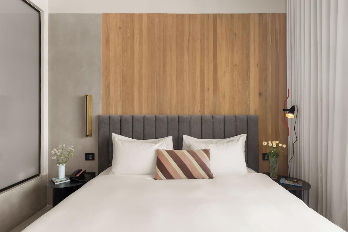 A modern hotel room with a large bed, a wooden accent wall, and a nightstand with a lamp and flowers.