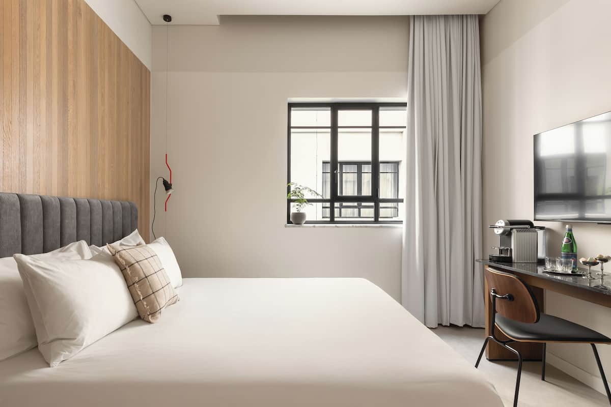 A modern hotel room with a large bed, a desk with a chair, a flat-screen TV, and a window with curtains.