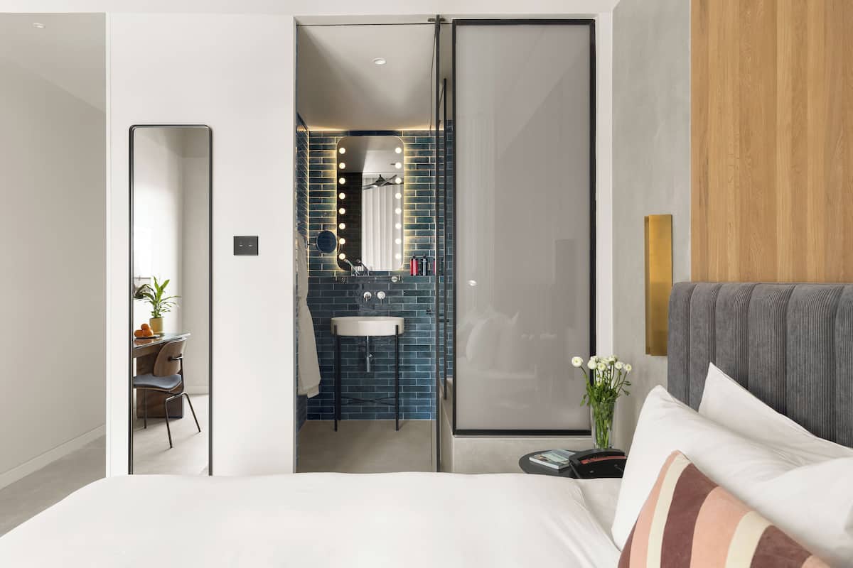 A modern hotel room with a large bed, a bedside table, a mirror with lights, a sink, and a wooden panel on the wall.