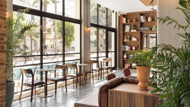 A cafe with large windows, wooden furniture, and a bookshelf. - Hotel Bazaar - Fattal Colors (Tel Aviv)