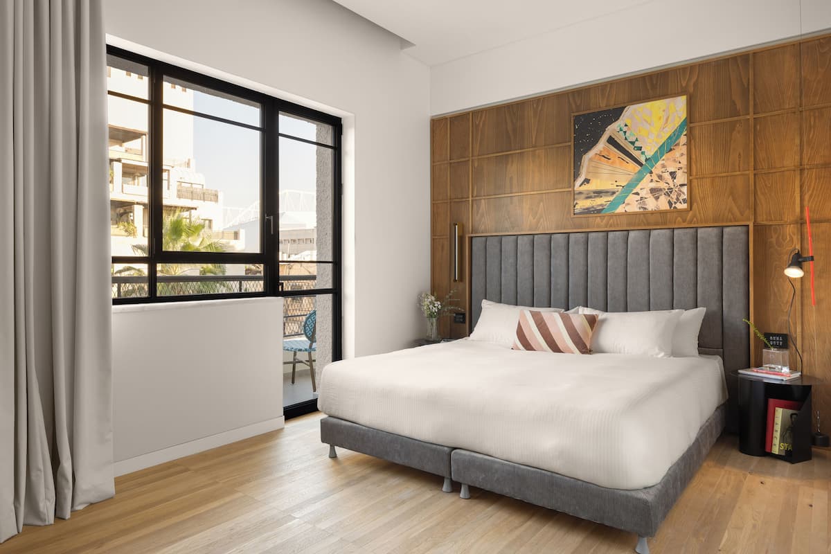 A modern bedroom with a large bed, a wooden headboard, a city view through a window, and a small side table with a lamp.